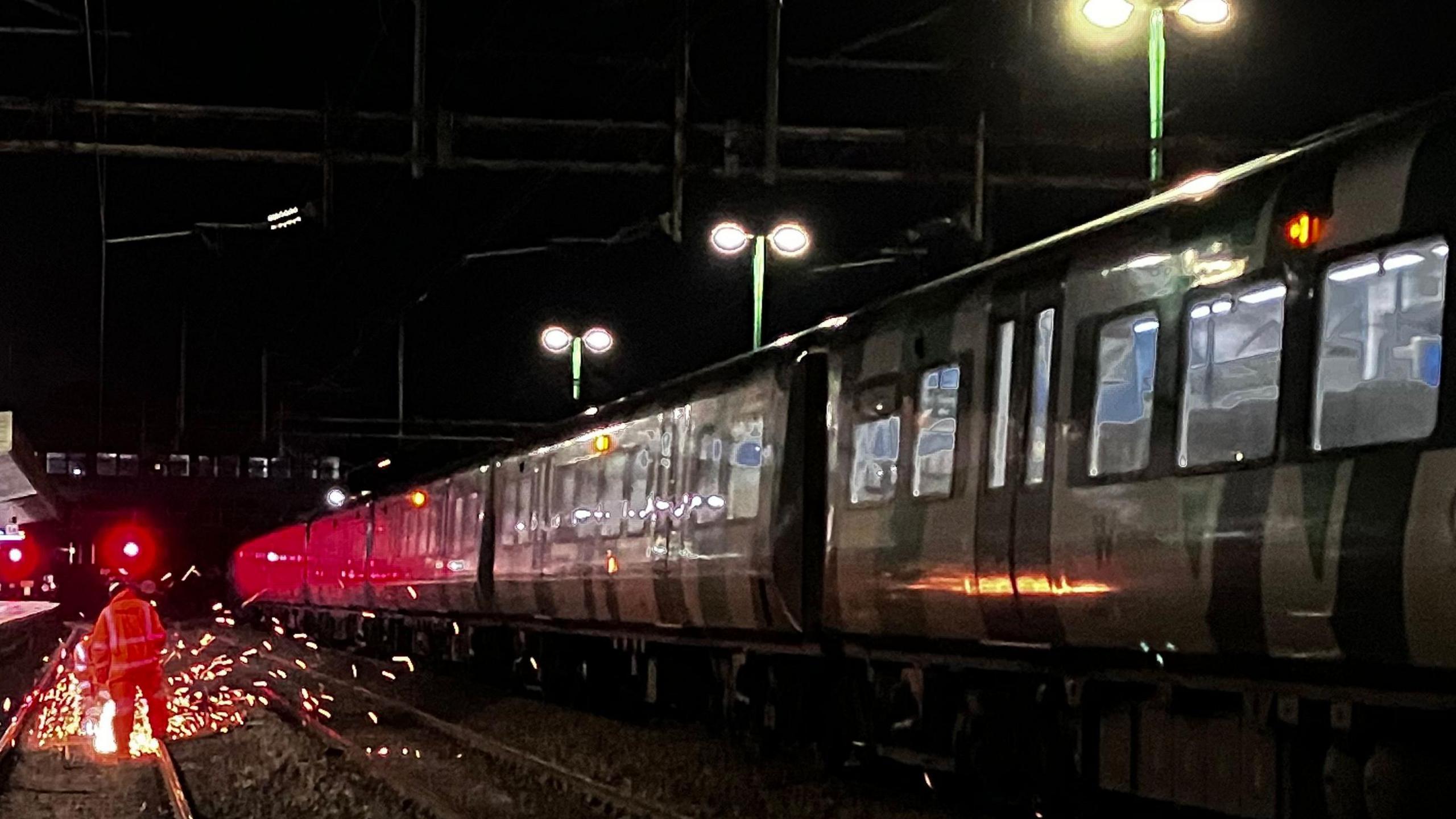 A multi-carriage train passes a man in a orange high visibility suit working on the train tracks. Sparks are coming off the tracks where the man is fixing the metal. 