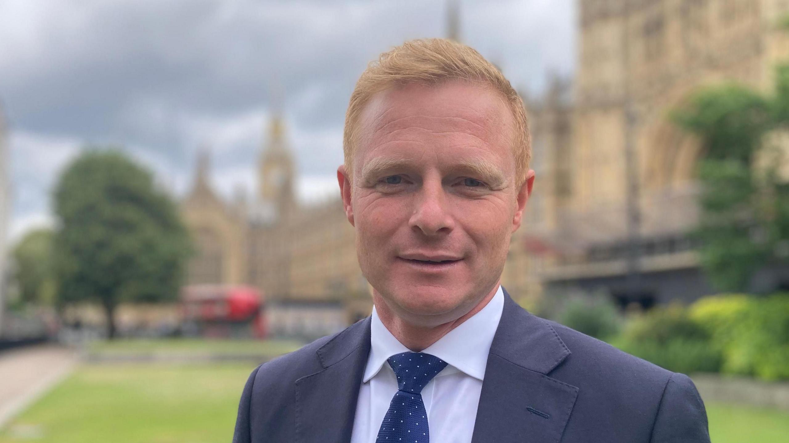 Robbie Moore, who has short ginger hair. He is standing outside the Houses of Parliament and wearing a blue suit with a white shirt and blue tie.