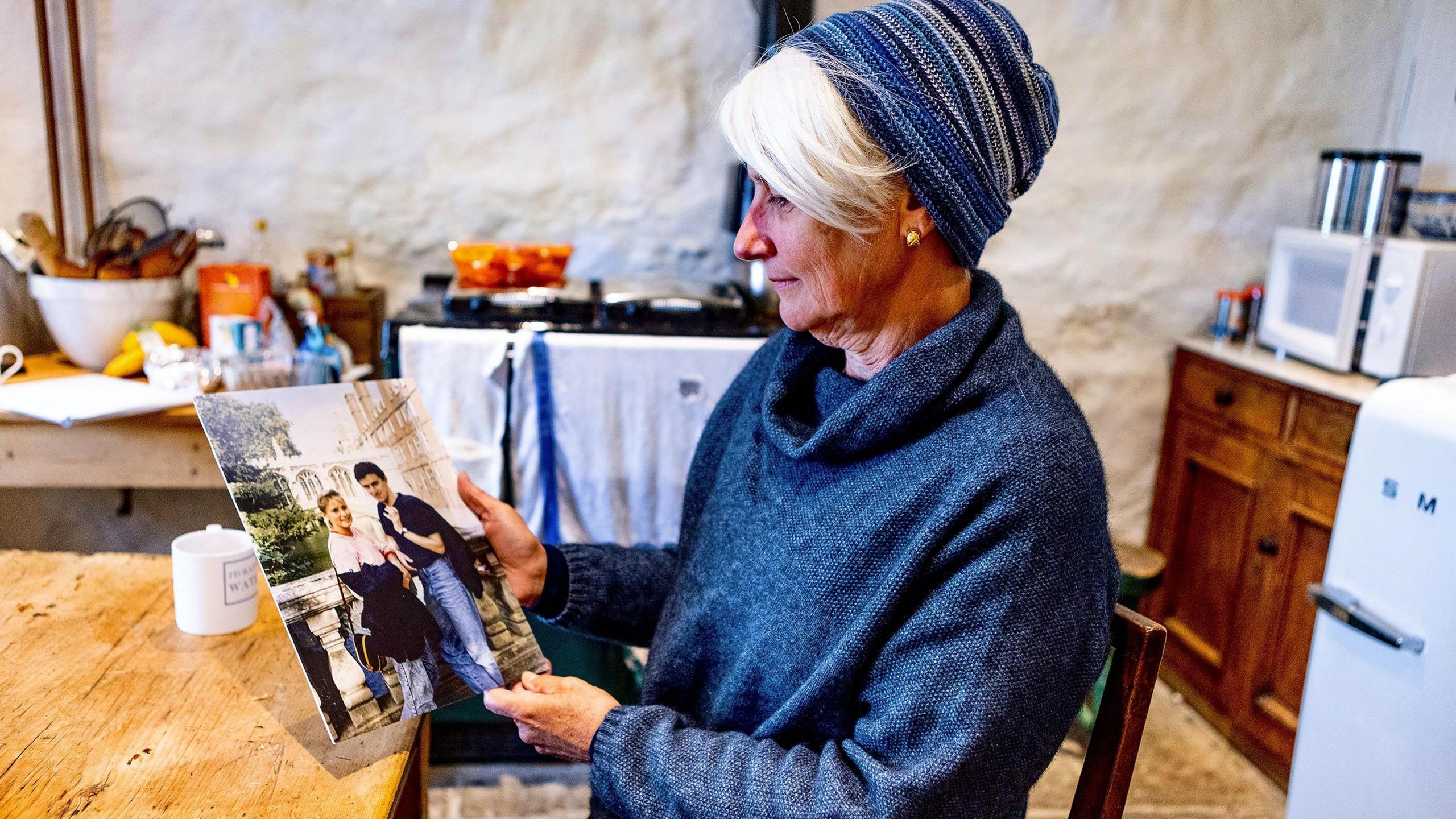 Rose Grant, now older wearing a blue jumper and hat, is sat at a kitchen table holding a photo of her with Tim when she was younger