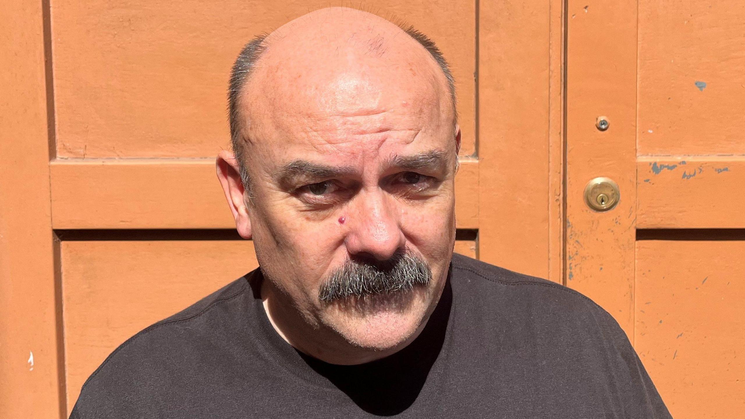 Paul Garrett with a moustache and black t-shirt against an orange door on a sunny day