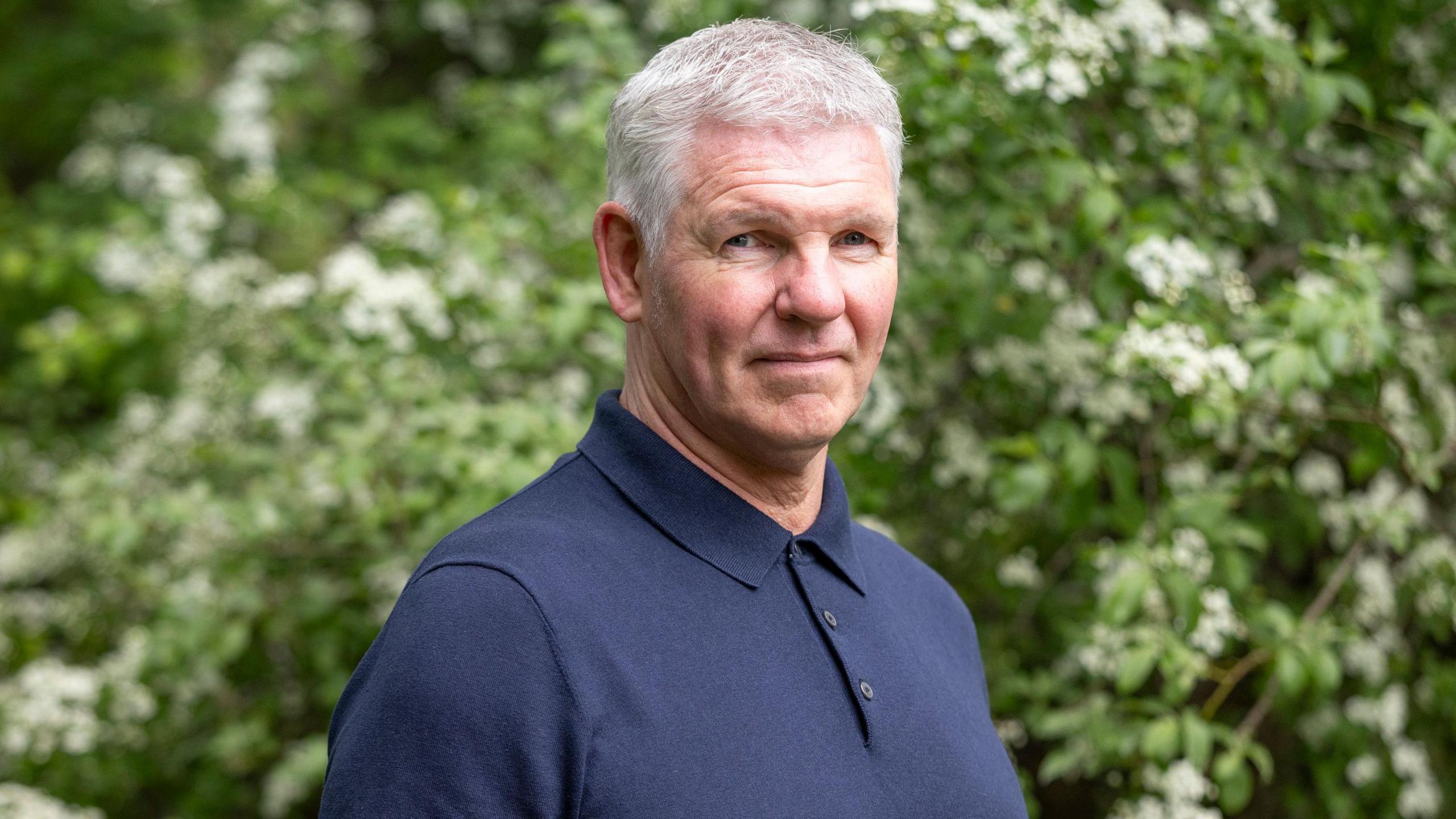 A man with grey hair and a dark blue t-shirt looks at the camera
