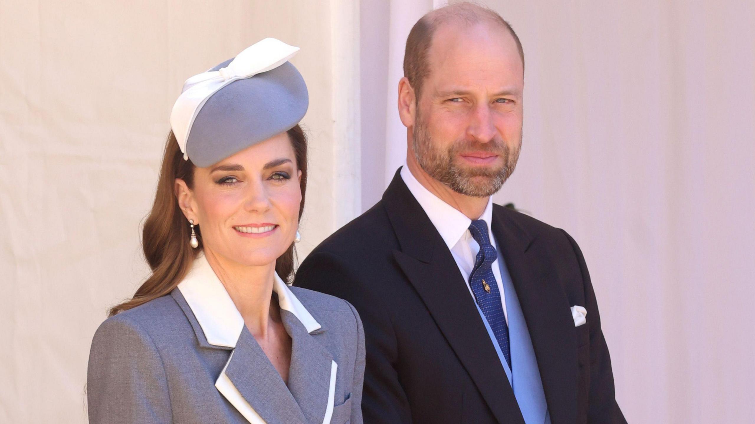 Princesha e Uellsit e veshur me një fustan gri-xhaketë me detaje të bardha në jakë dhe një kapelë gri dhe të bardhë - Princi i Uellsit ka një kostum të errët me jelek blu të zbehtë dhe një kravatë blu të errët.