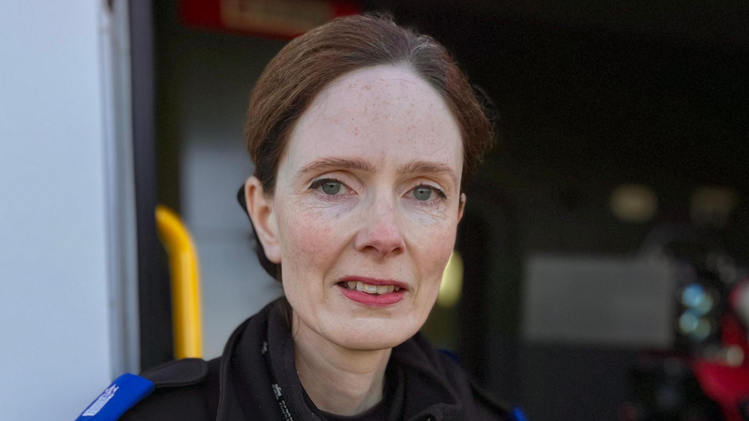 Cara Taylor smiles at the camera. She has dark red hair that has been tied back in a bun behind her head. She stands in front of police speed enforcement van that has its doors open offering a view inside.