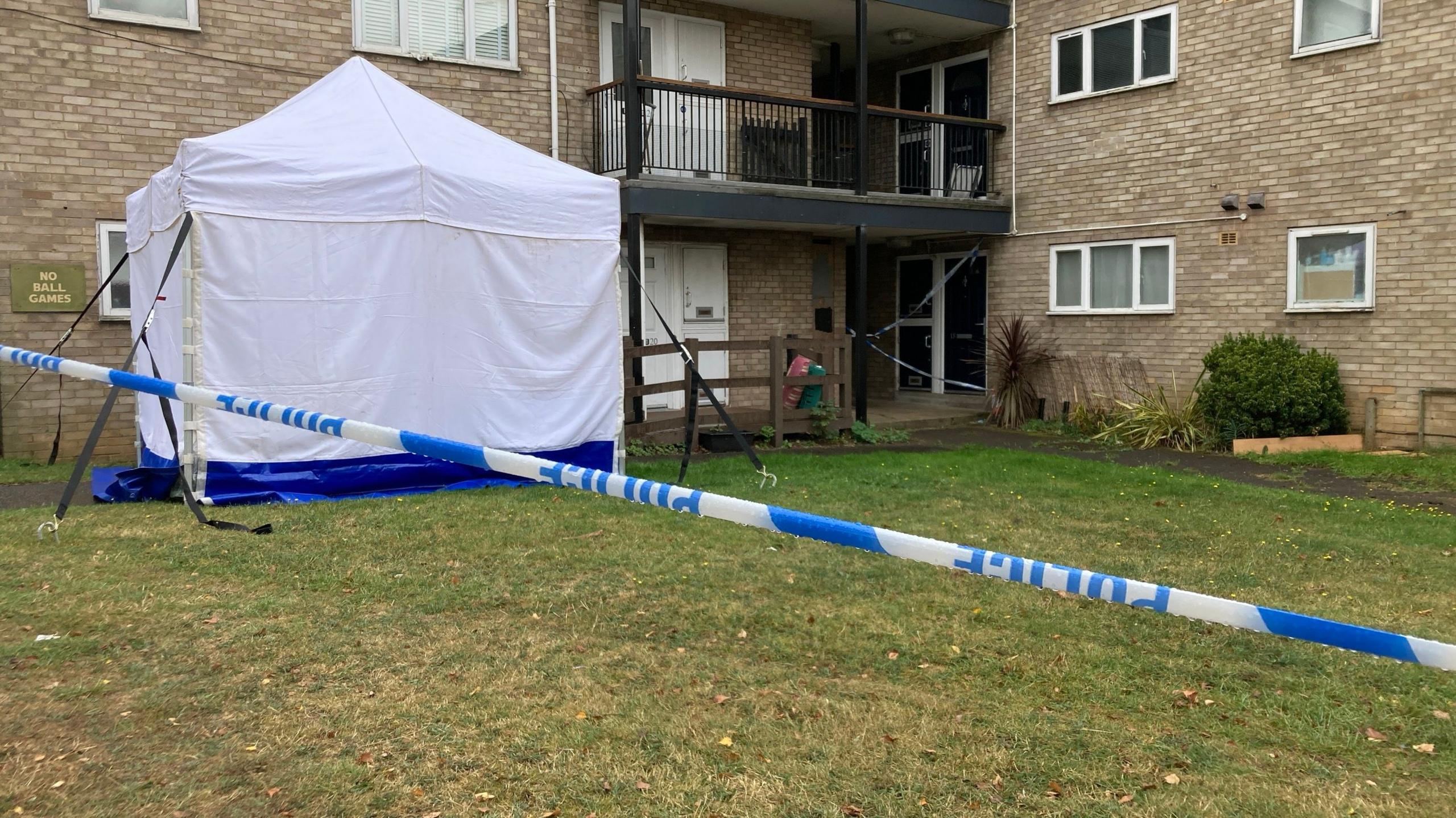 A large white forensic tent outside a block of flats with police blue and white tape in the foreground.