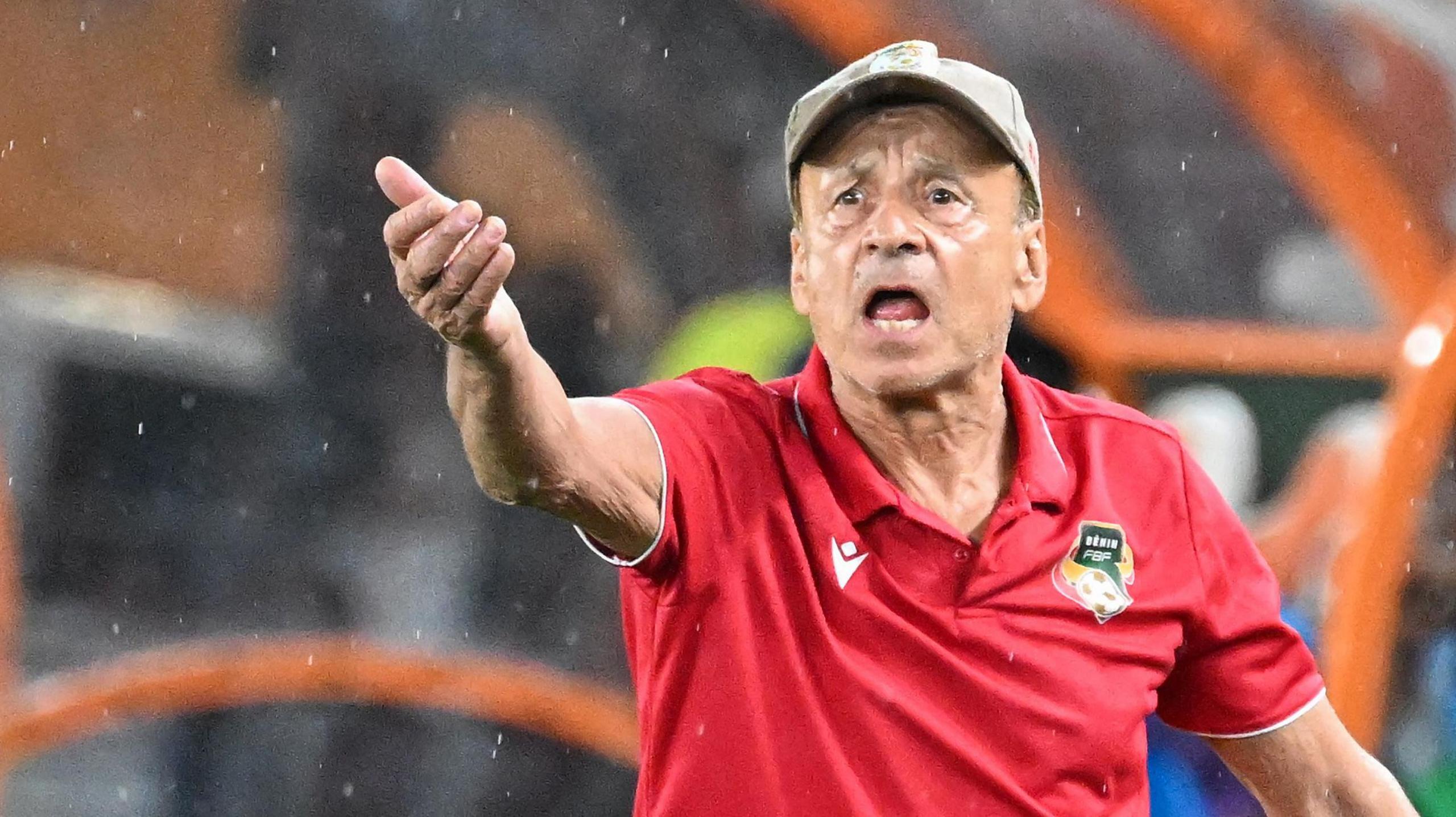 Gernot Rohr wears a beige cap and red polo shirt with Benin football crest on it as he raises his right arm in front of him and gesticulates on the touchline with a look of angst on his face