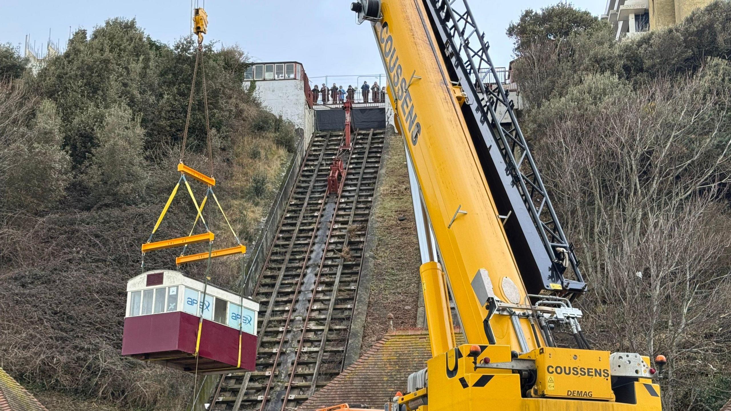 A funicular with a red carriage being lifted off the track by a yellow crane. At the top of the funicular is a group of people stood watching the removal. 