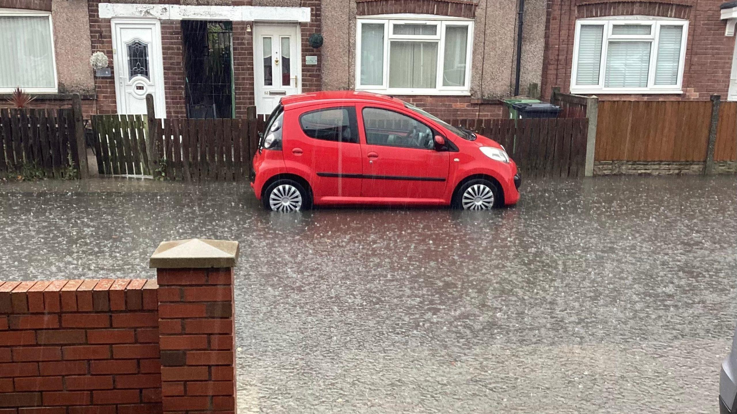 A bright red car is parked on a street with rain flooding the road.