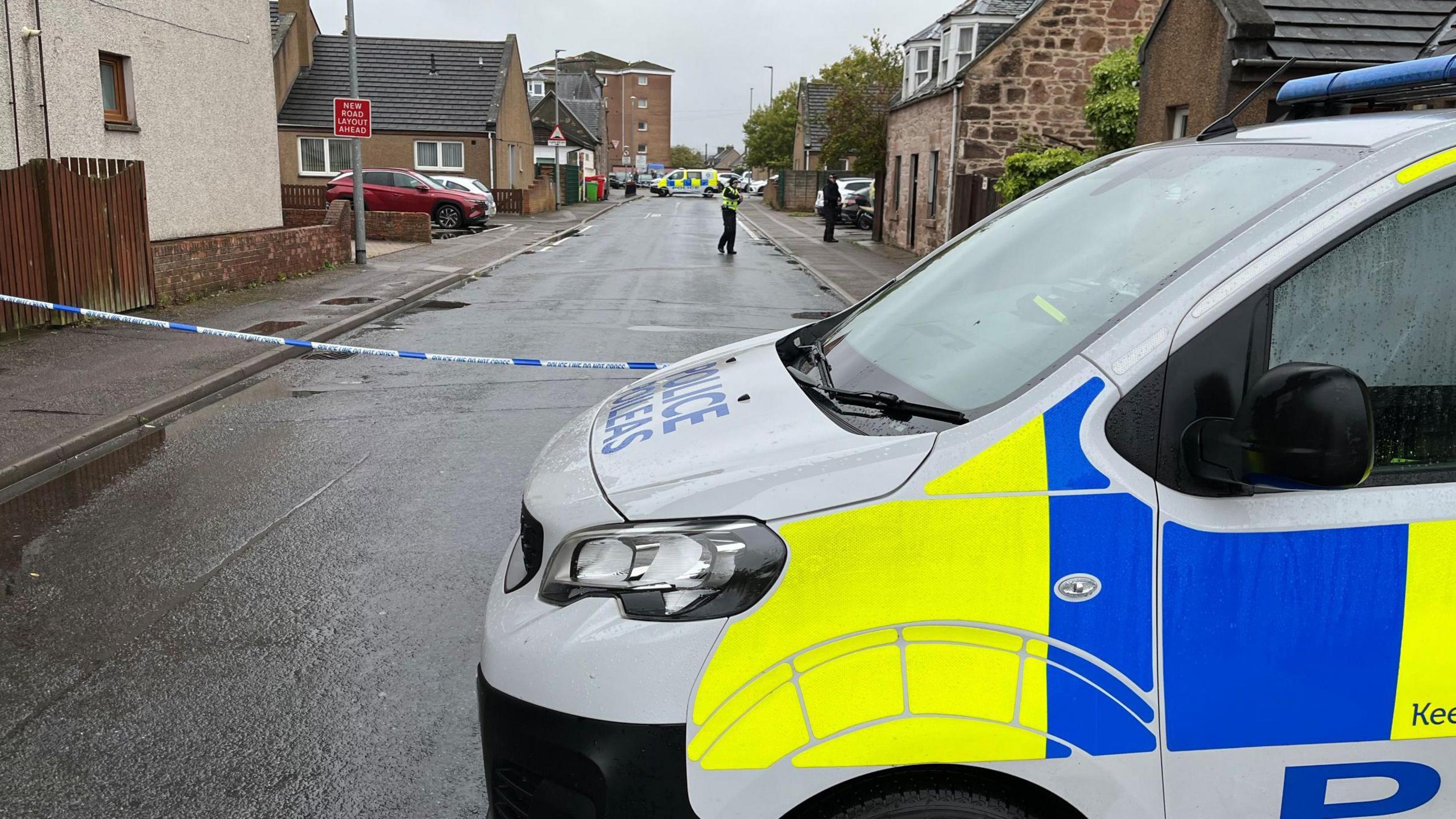 A partial side view of a police van parked in front of a street in Merkinch. The street is lined by different types of properties, including traditional stone-built houses and more modern blocks of flats. The street is wet following a rain shower, and there is police tape stretched across the street next to the van. A police office and another police van are further up the street.