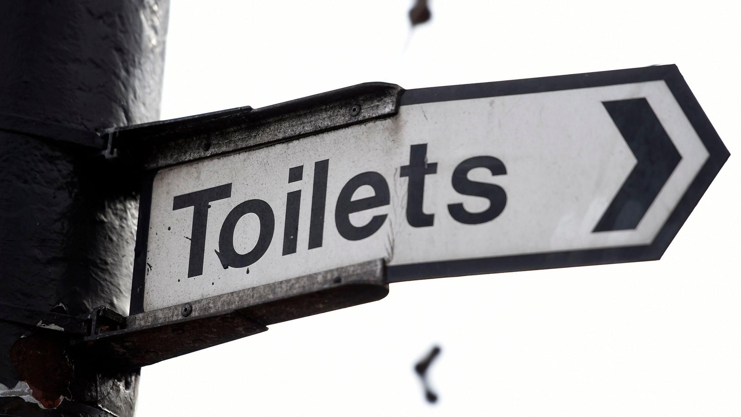 A black and white street sign points to the right, indicating the nearest toilets. The sign is a little bent in the middle, and it's attached to a post.