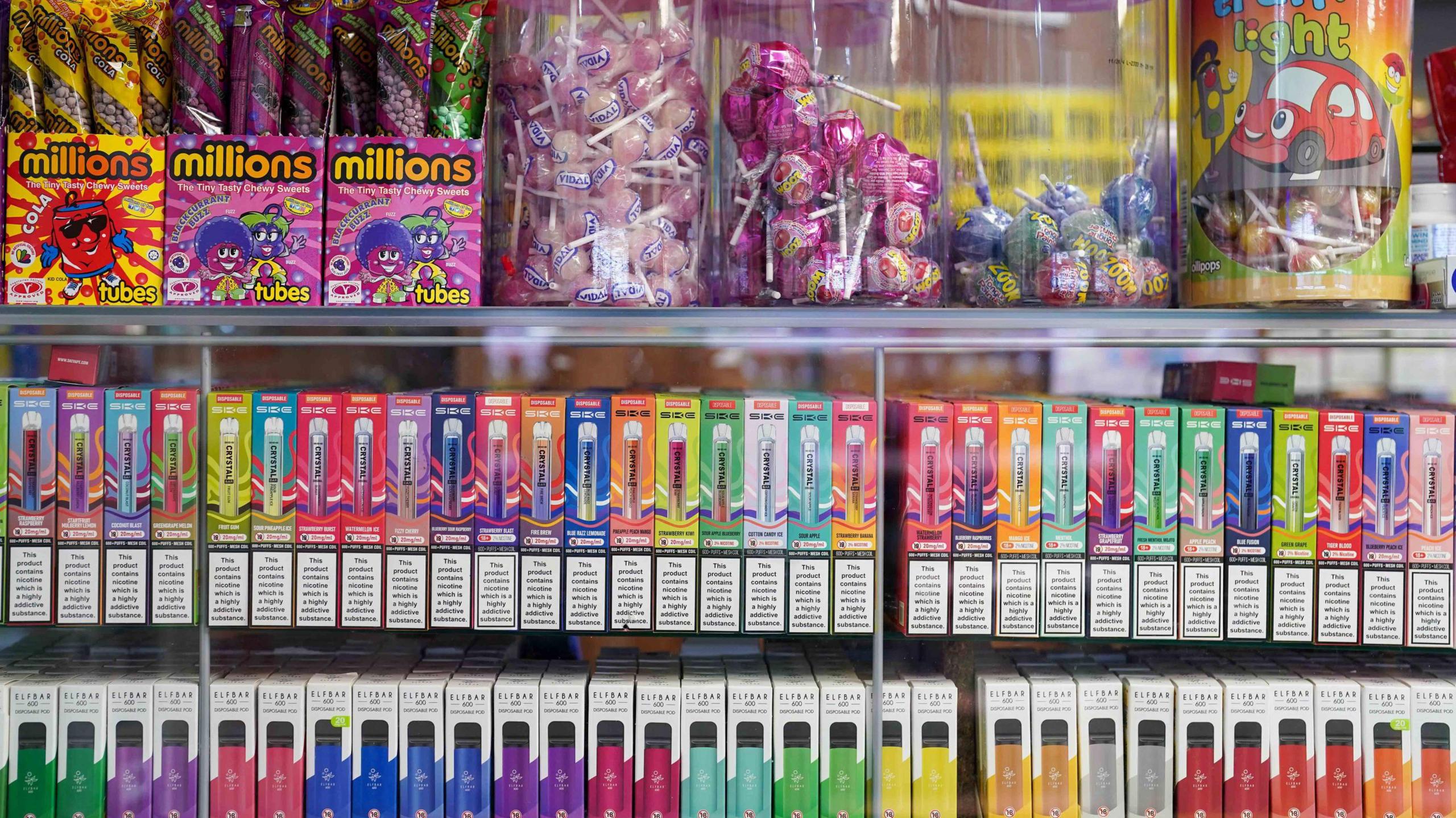 Disposable vapes of varying colours and flavours on display at a corner shop. There are sweets on display above the vapes.