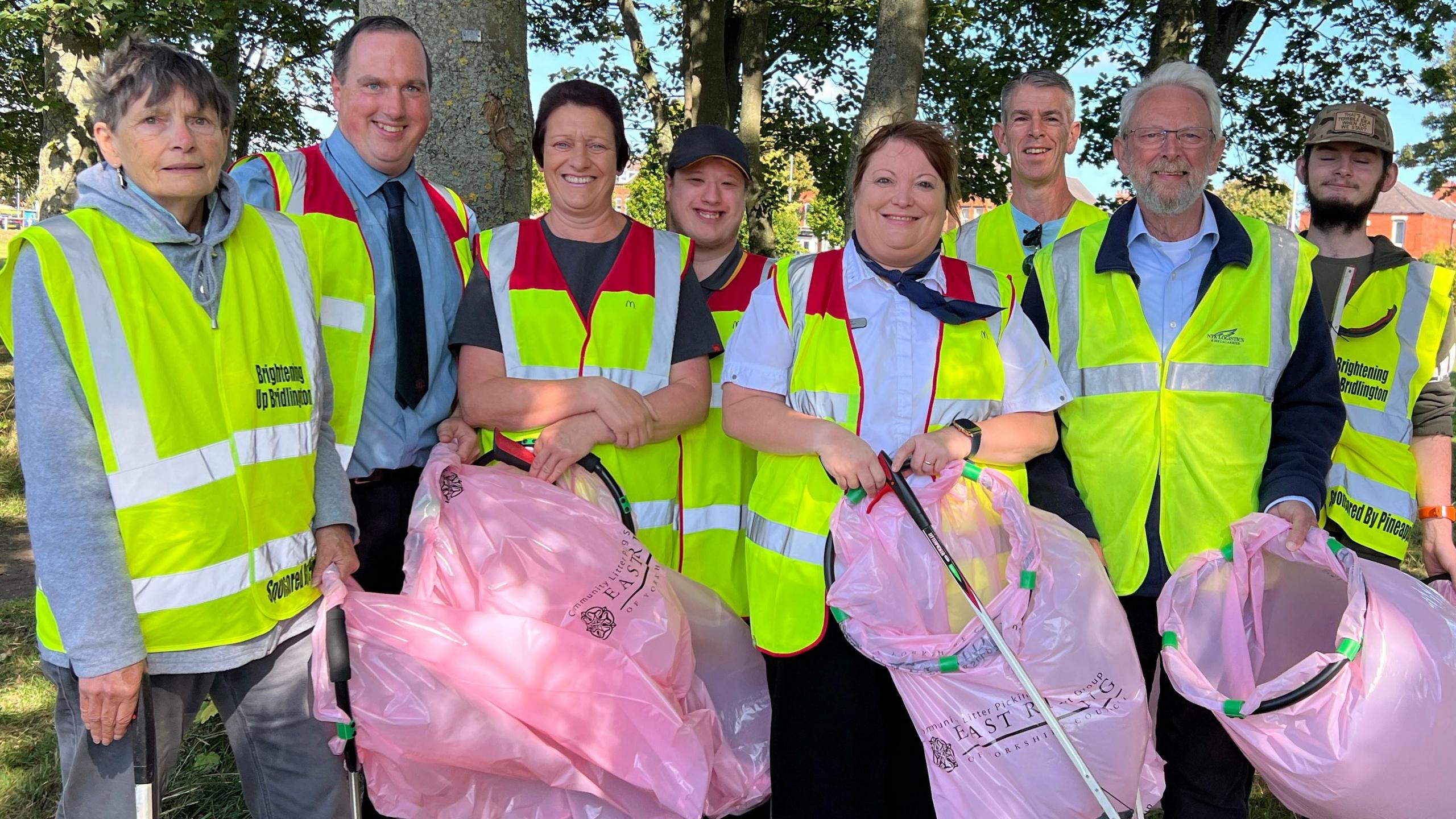 A group of eight men and women wearing high-visibility vests and carrying pink litter bags and long litter-picking grabbers. They are standing in a park in front of trees.