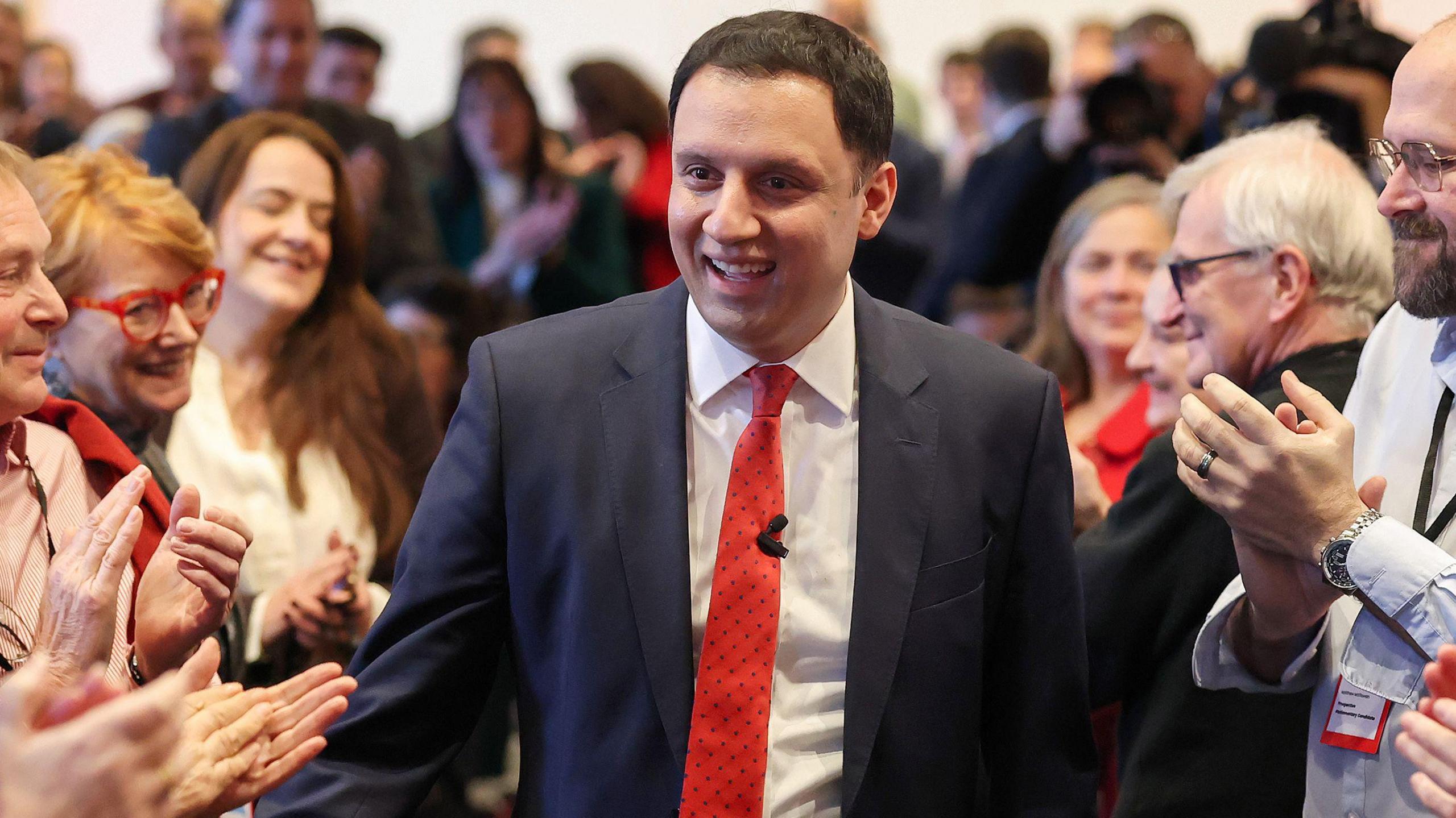 Anas Sarwar in a dark suit and red tie walking through a crowded room while being applauded by attendees on both sides