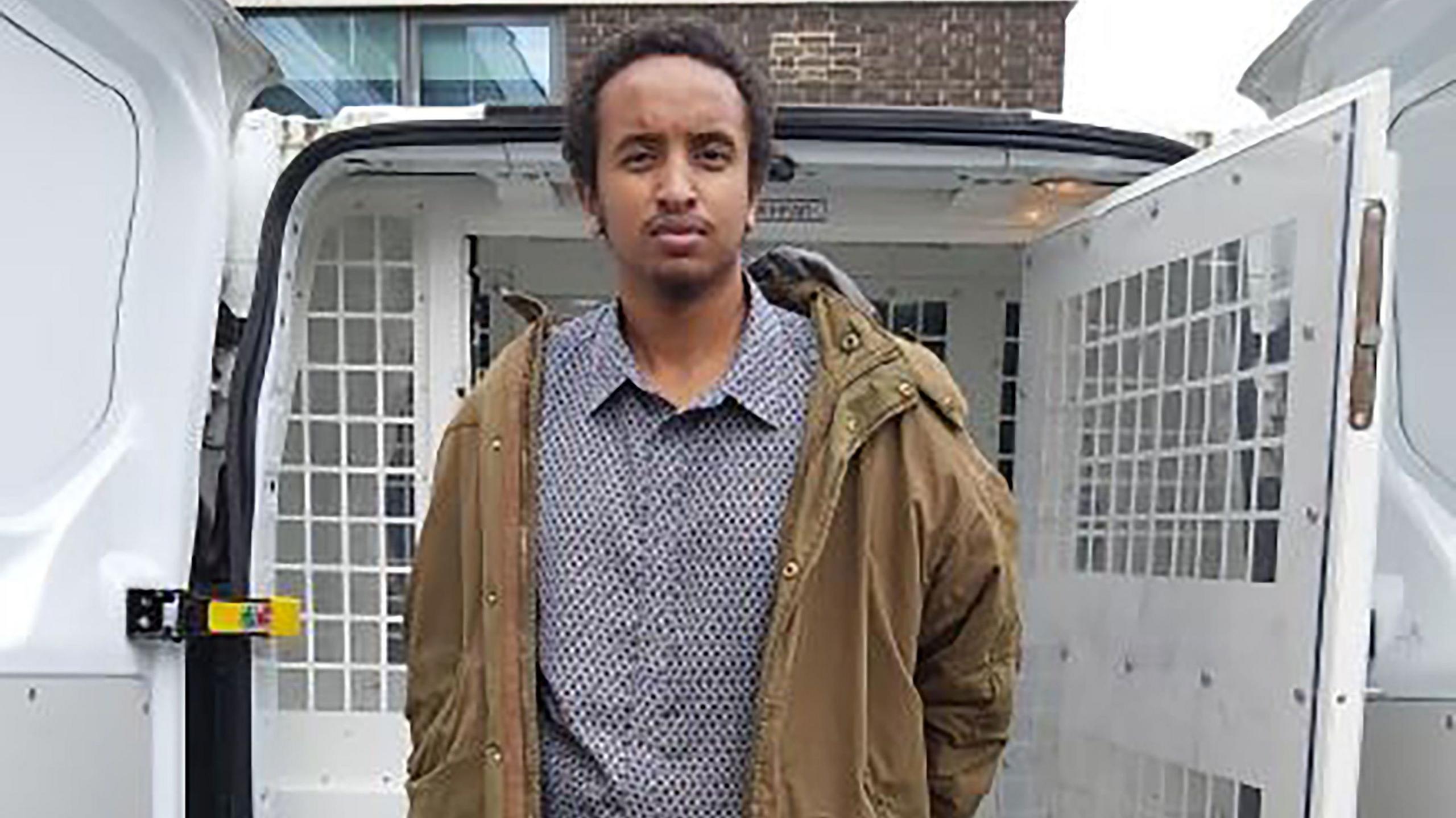 Ali Harbi Ali, a man wearing a grey shirt and brown coat, apparently has his hands handcuffed behind his back as he stands in front of a prison van.
