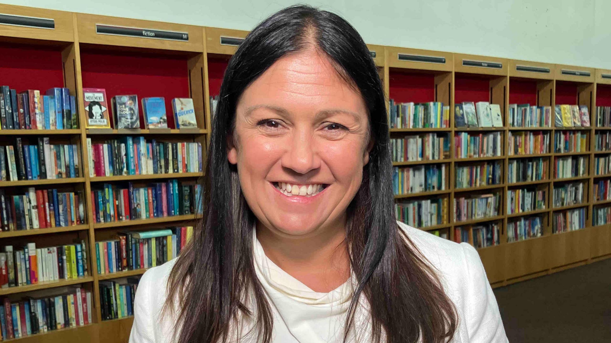 Lisa Nandy is smiling at the camera while standing inside a library. She has long brown hair and is wearing a white longs sleeved blouse. Behind her are rows of book shelves filled with books. 
