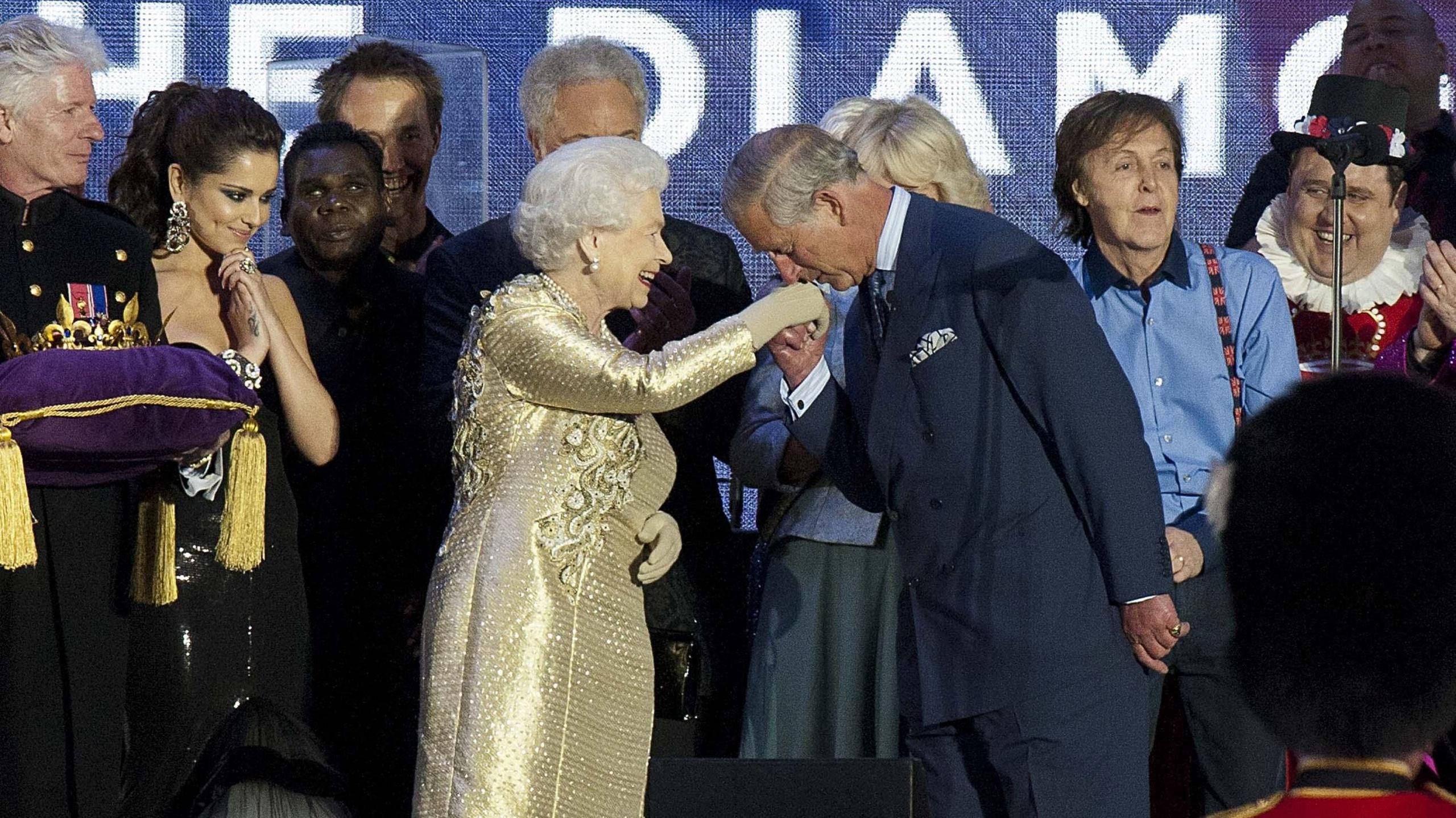 The Queen wears a gold dress as she stands on stage at her Diamond Jubilee concert. Her son Charles is kissing her hand as celebrities stand behind them smiling.