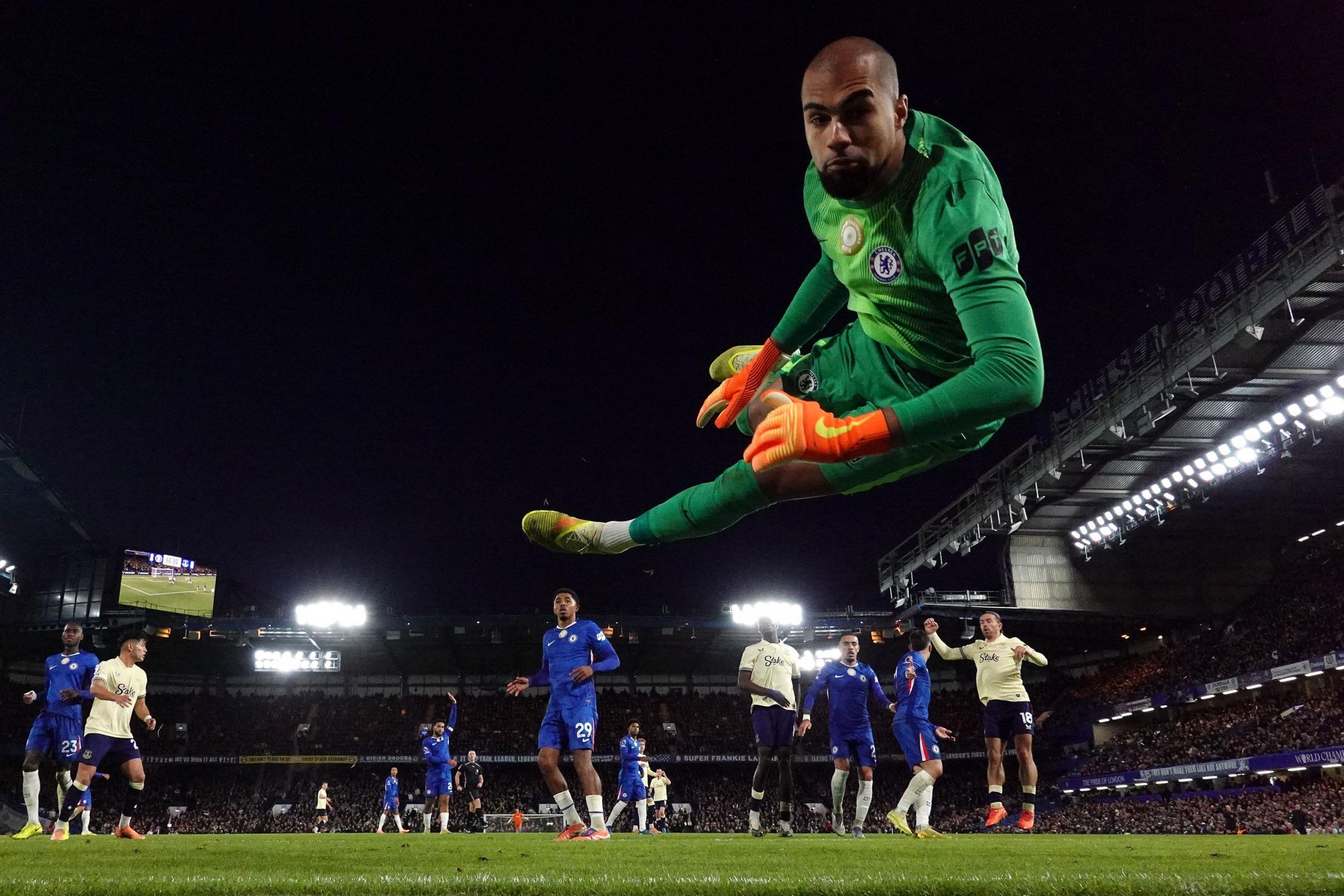 Un portero con un uniforme verde salta horizontalmente en el aire con las piernas completamente extendidas mientras intenta realizar una atajada durante un partido de fútbol nocturno, mientras los jugadores de ambos equipos observan cómo se desarrolla la acción dentro de un estadio brillantemente iluminado.