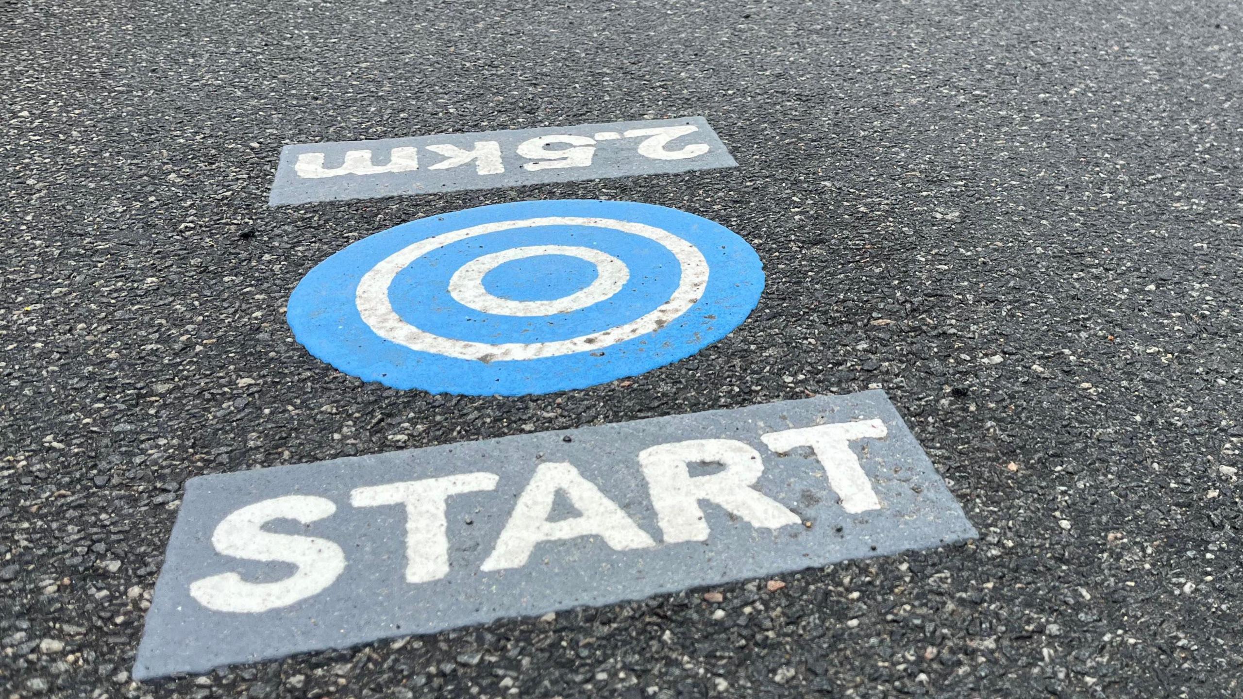 There are three parts to the marker which is on a tarmac path. The middle bit has five blue and white concentric circles, one block to the left of it reads: 2.5km and the other to the right reads: START.