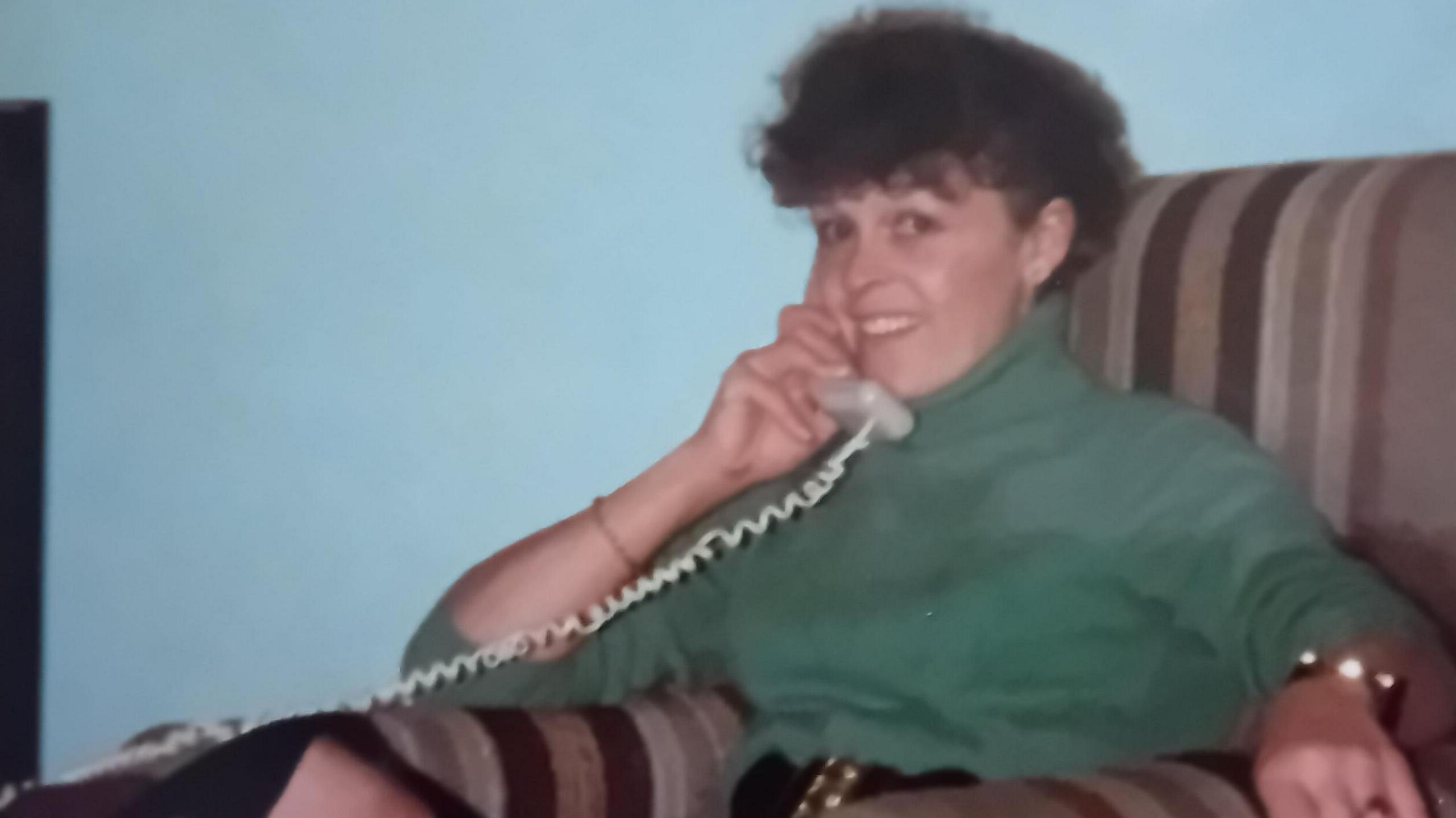 An old colour photo of a woman sitting on an armchair speaking on the phone.