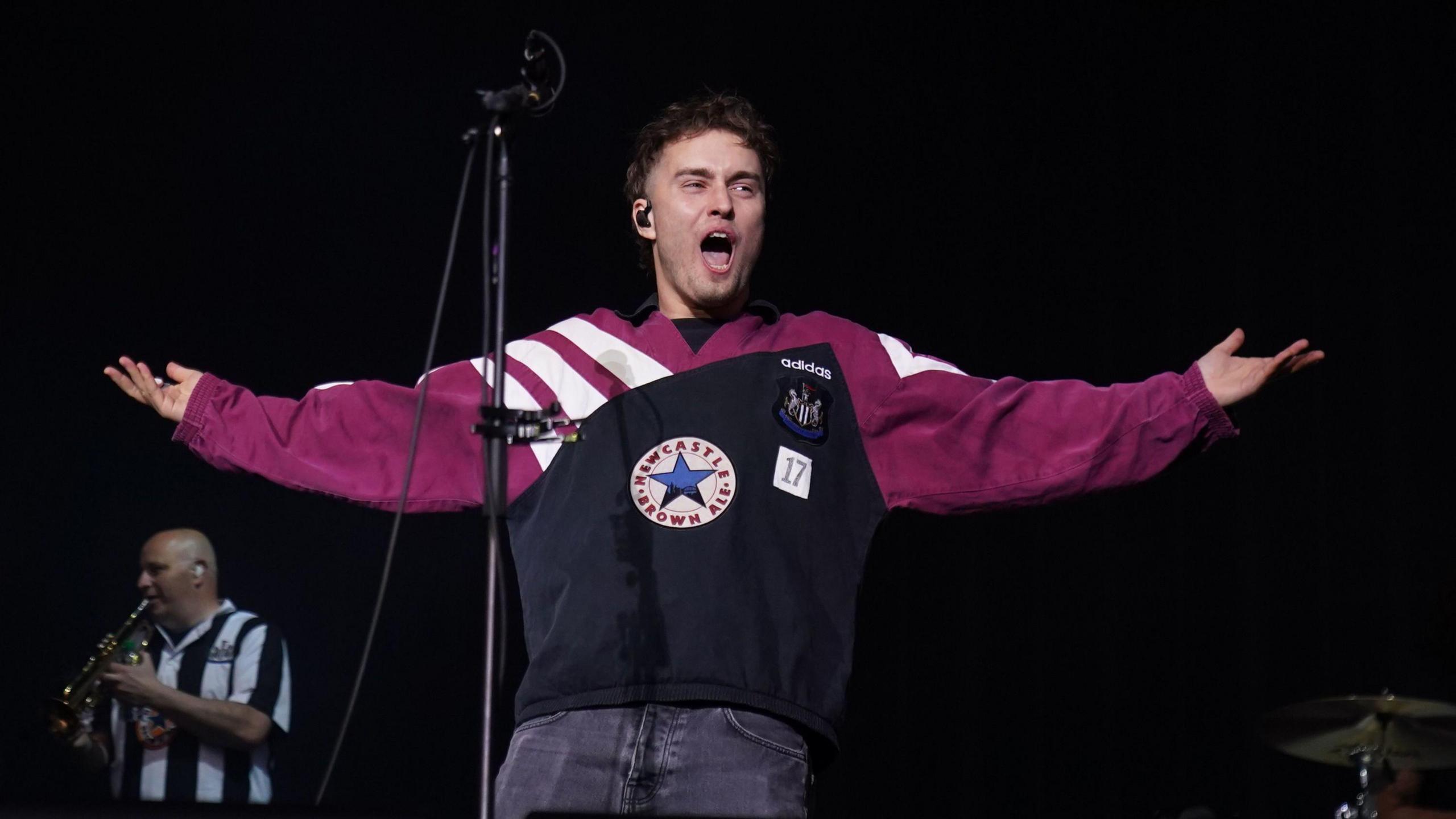 Sam Fender on stage with his arms outstretched, shouting. He's wearing a black and maroon vintage Newcastle United top.