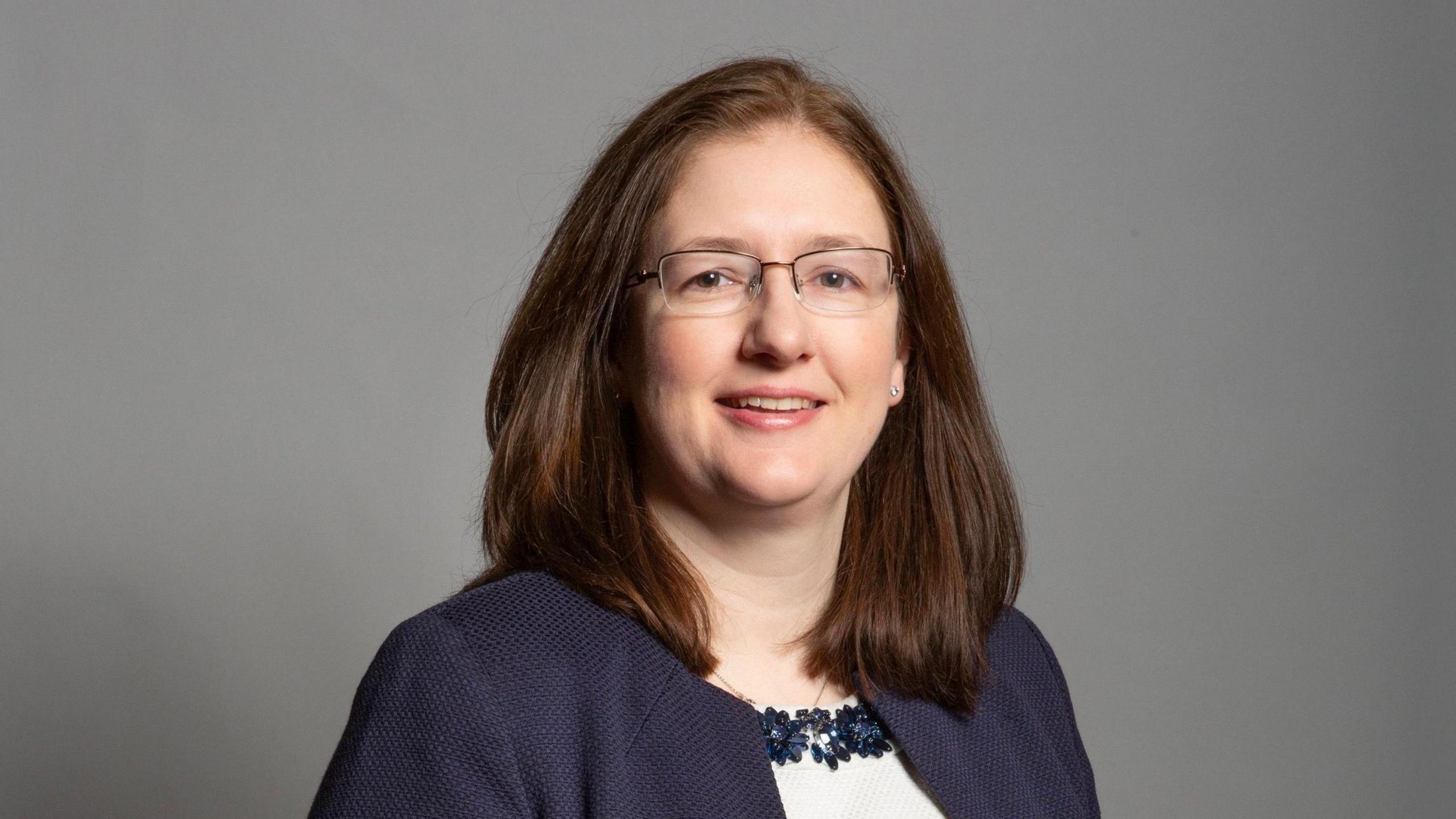 Head and shoulders shot of Dr Caroline Johnson. She has dark shoulder-length hair and is wearing glasses and a blue jacket.