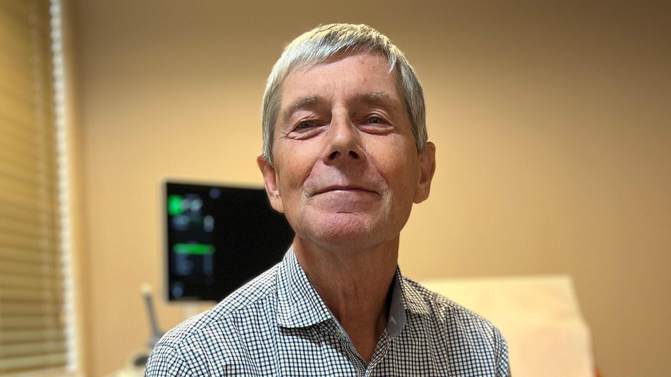 Neil looks at the camera as he sits on a white leather chair in a medical consultation room. He has a thin striped shirt on and behind him is a reclining chair next to a medical screen. Neil has short grey hair.