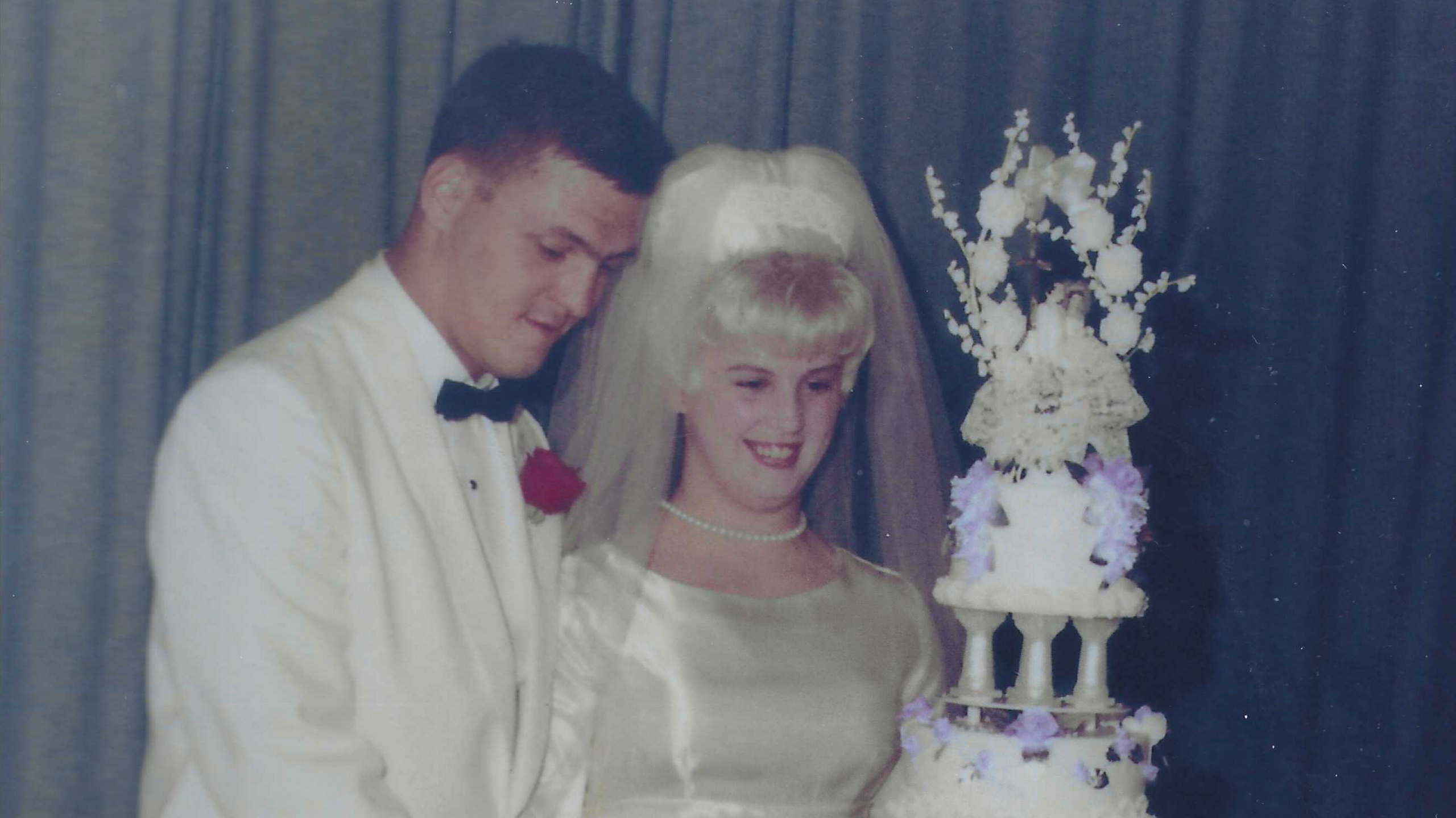 An old faded colour photo of a man in a white suit and black bow tie and a smiling woman wearing a white wedding dress and veil. They are standing next to an elaborate multi-tiered wedding cake.