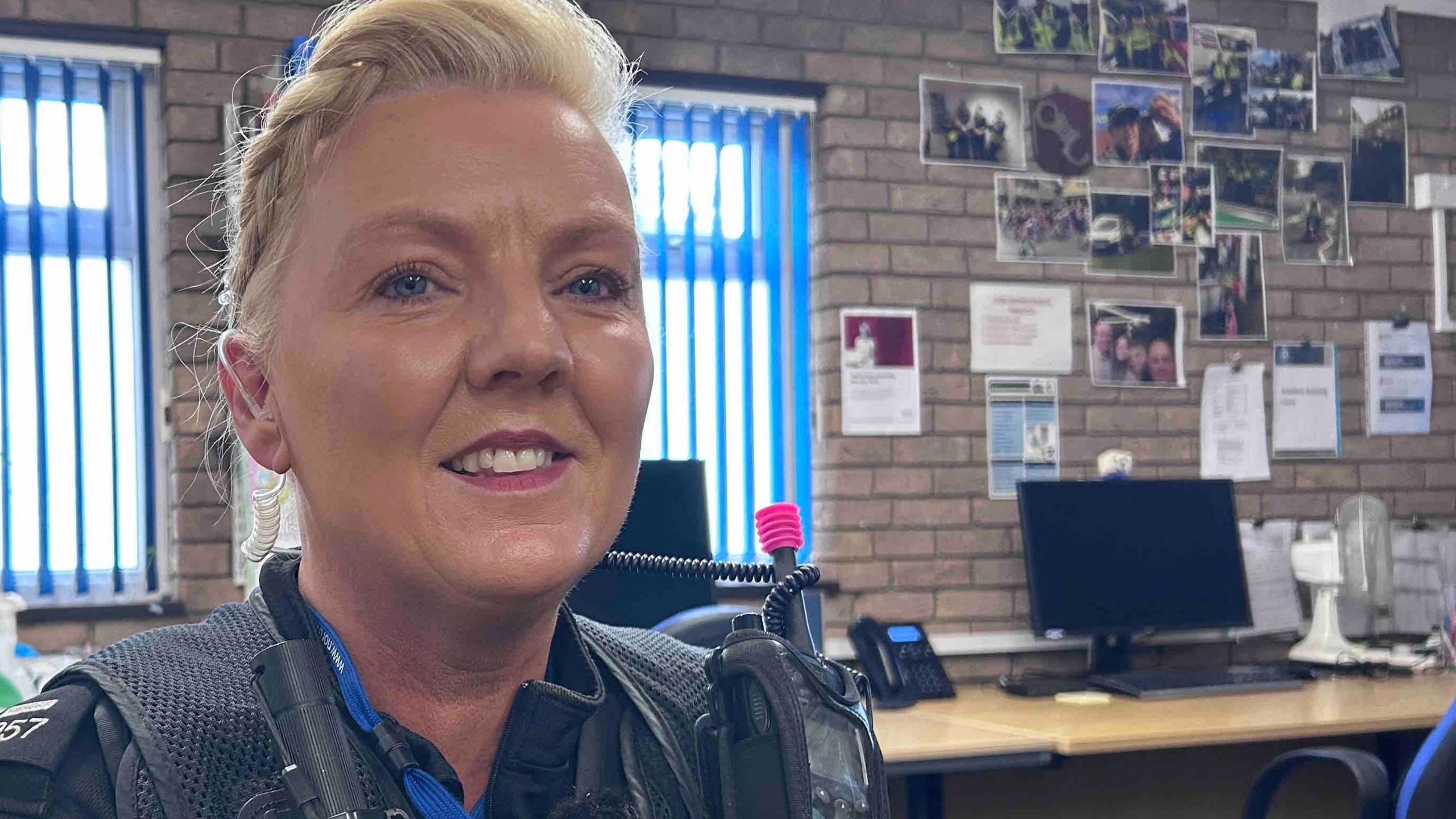 Lorna Montecalvo is sitting inside an office wearing her police uniform. She is looking directly at the camera and smiling and has blonde hair which is braided back.