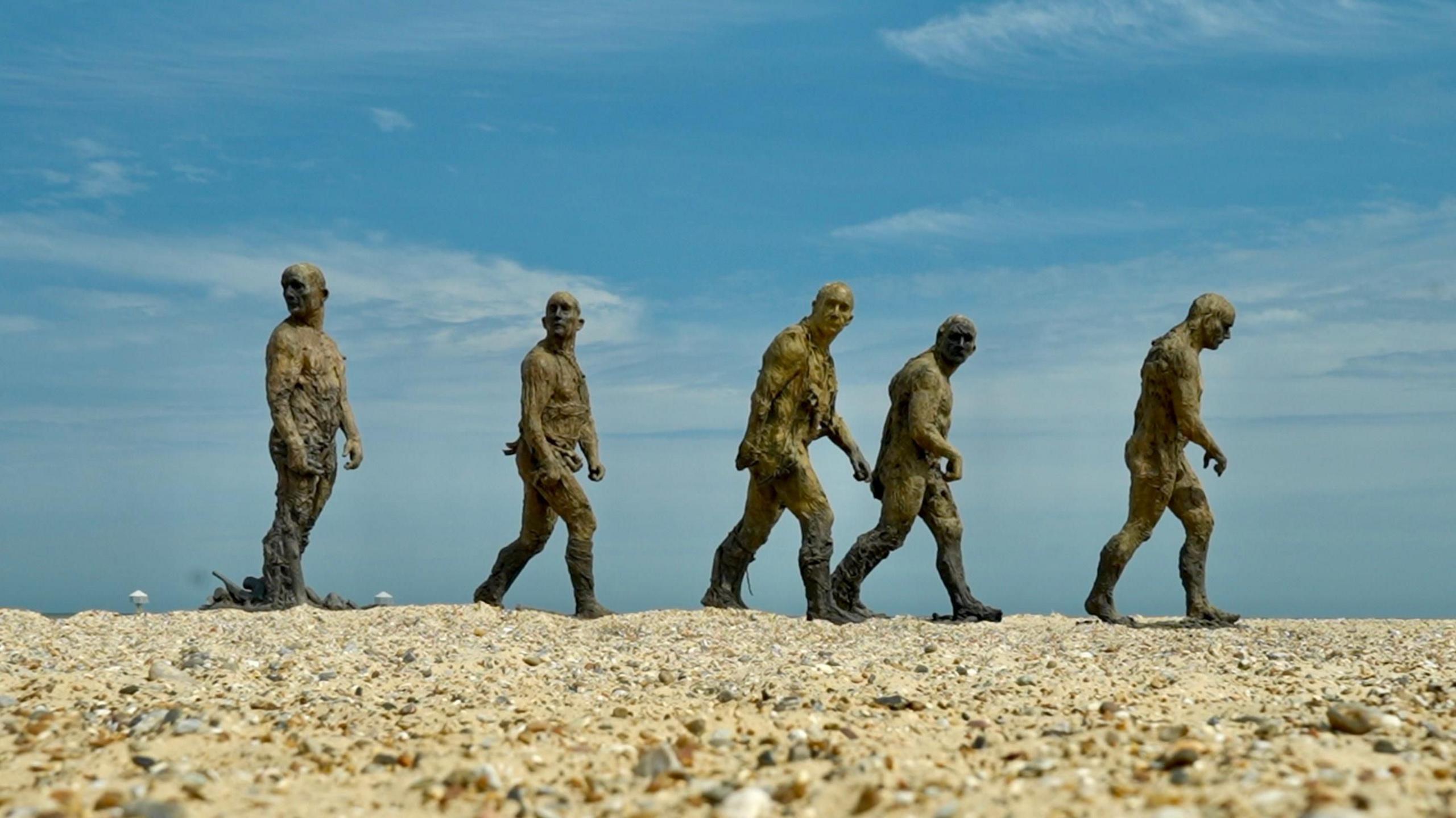 Five bronze men in walking poses at a beach.  Most are slightly stooped. Three are looking towards the beach, the other two are looking forwards.