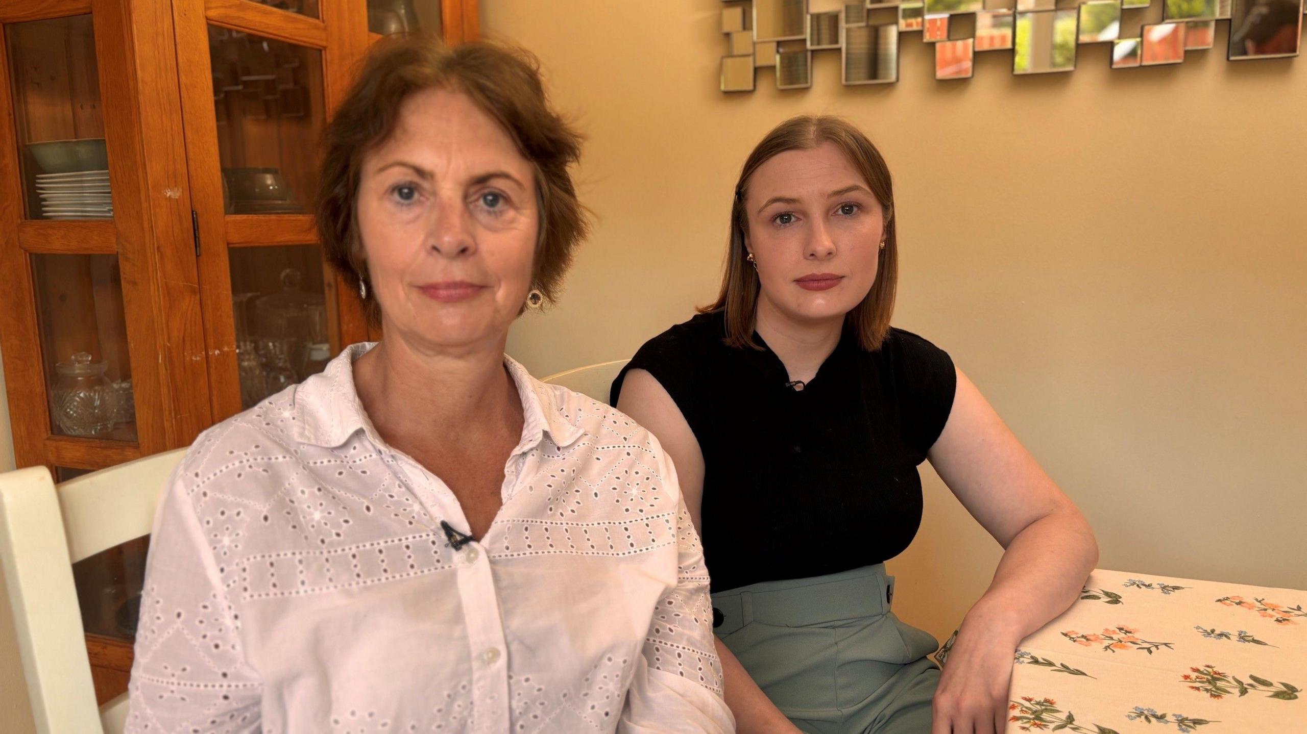 Susan and Anna - Susan sits on the left and wears a white shirt. Sh4e has brown hair and a neutral expression. 