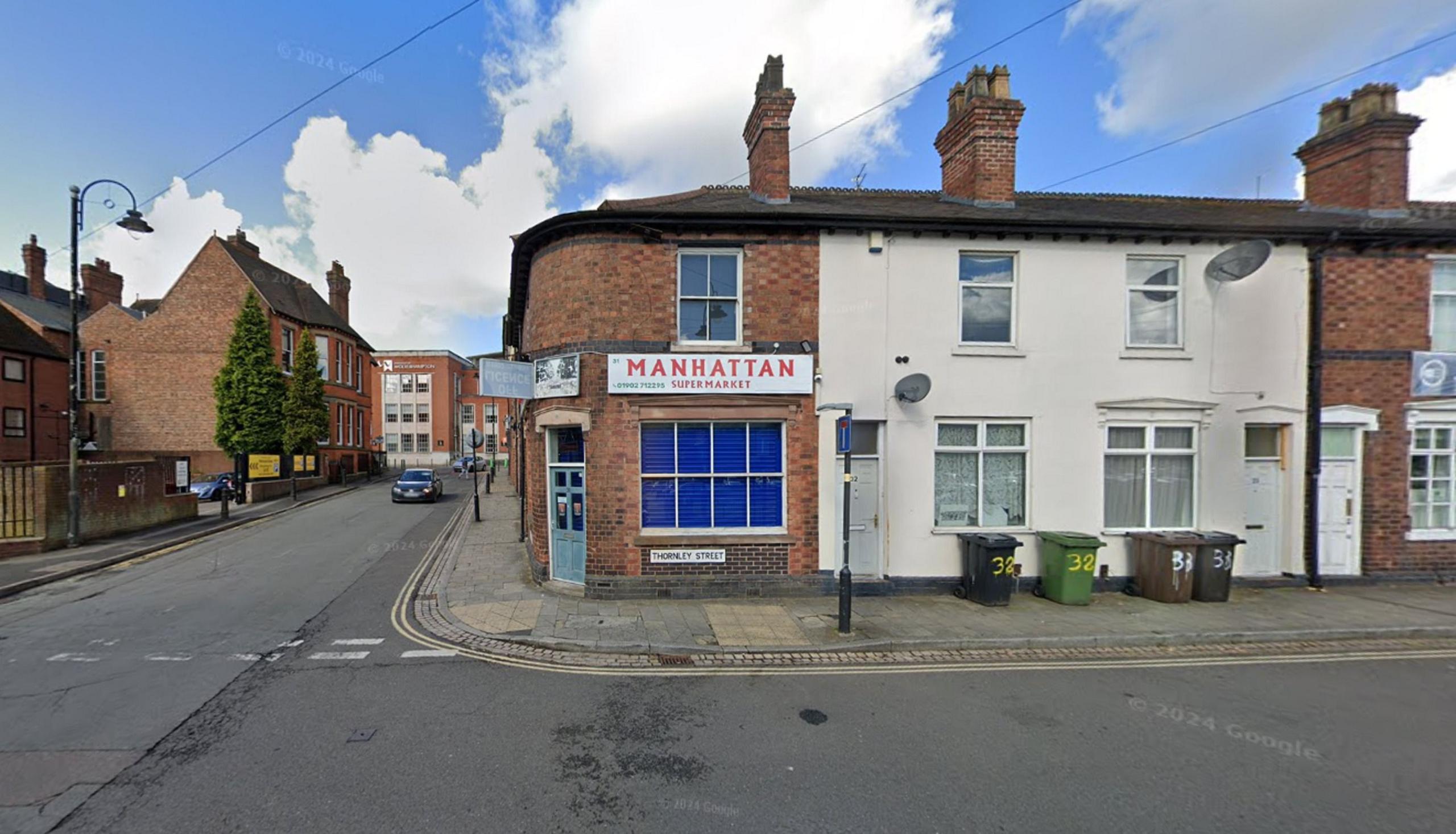 A corner shop, the building is a red-brick terraced house, with a white sign and red lettering saying Manhattan in capital letters
The door is light blue and the windows have dark blue shutters 