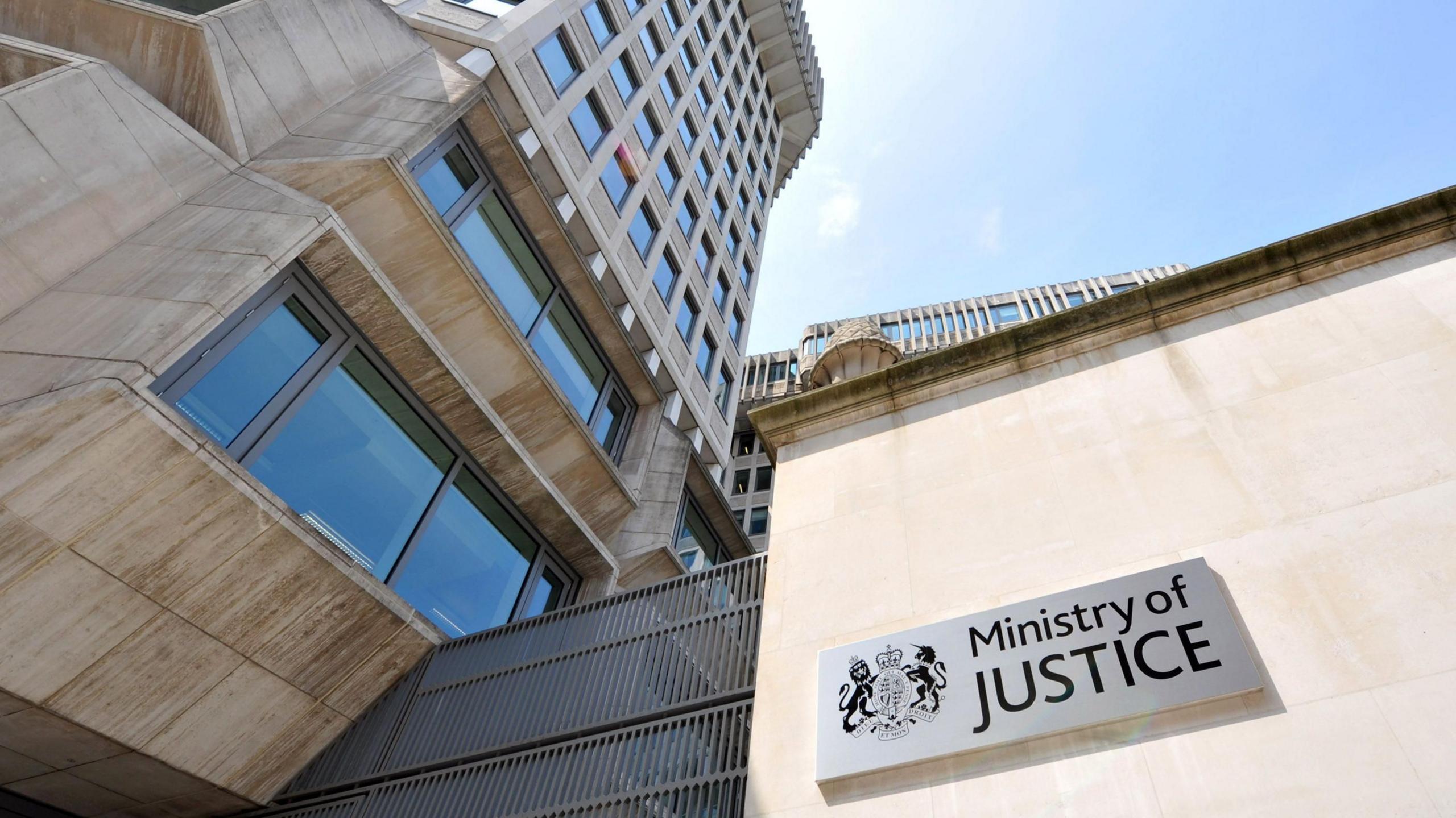 A Ministry of Justice building with a sign saying Ministry of Justice against a stone clad wall with a concrete tower block reaching to the left and pale blue sky above