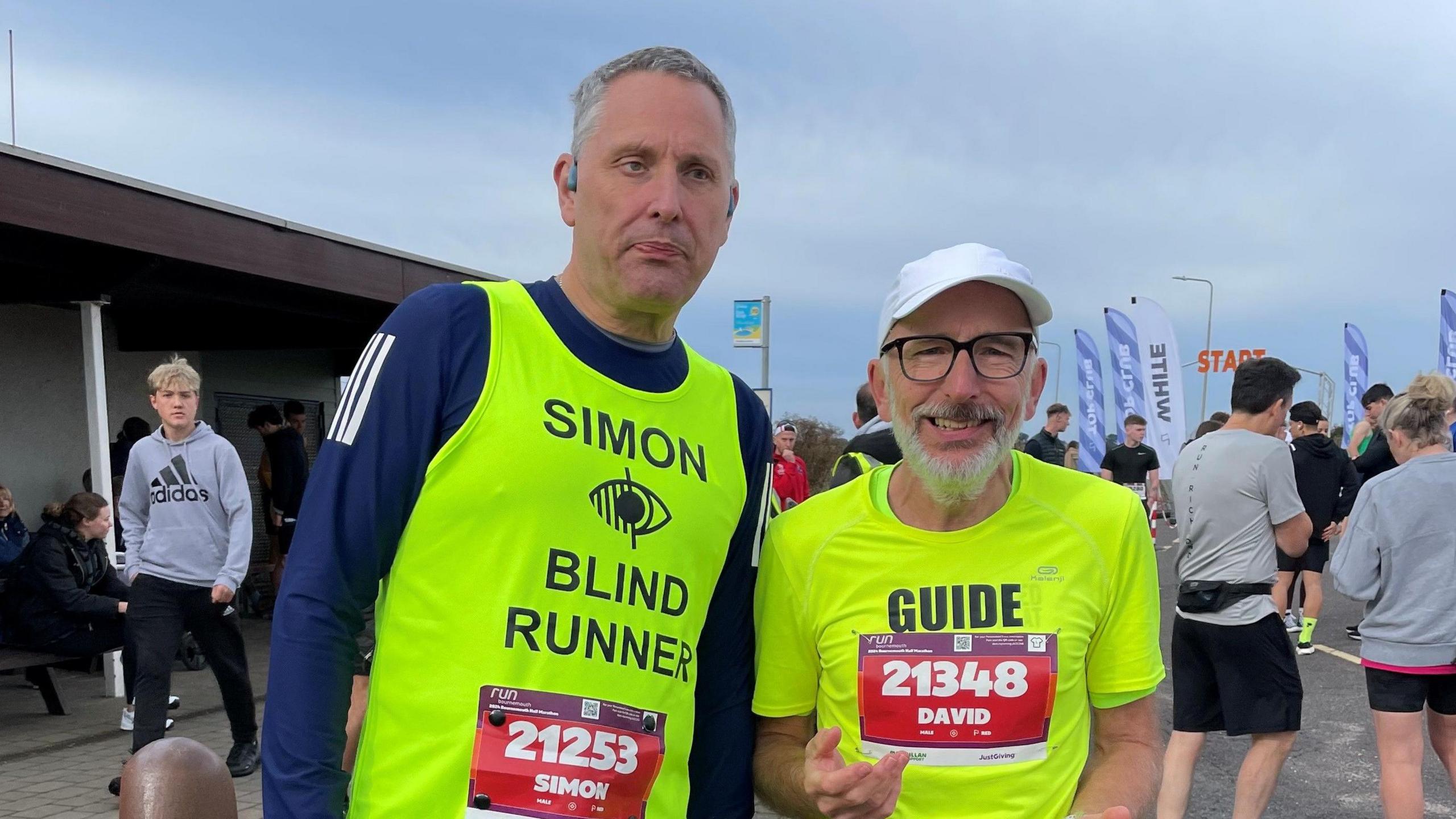 Simon Goodall, a tall, white man with grey hair. He is wearing a dark blue running top with a high-vis vest over it which his name and reads 'BLIND RUNNER'. He is standing next to David Green, a middle-aged white man of a small build, with a grey beard and moustache. He is wearing a white cap and a bright yellow high-vis t-shirt which reads 'GUIDE'.