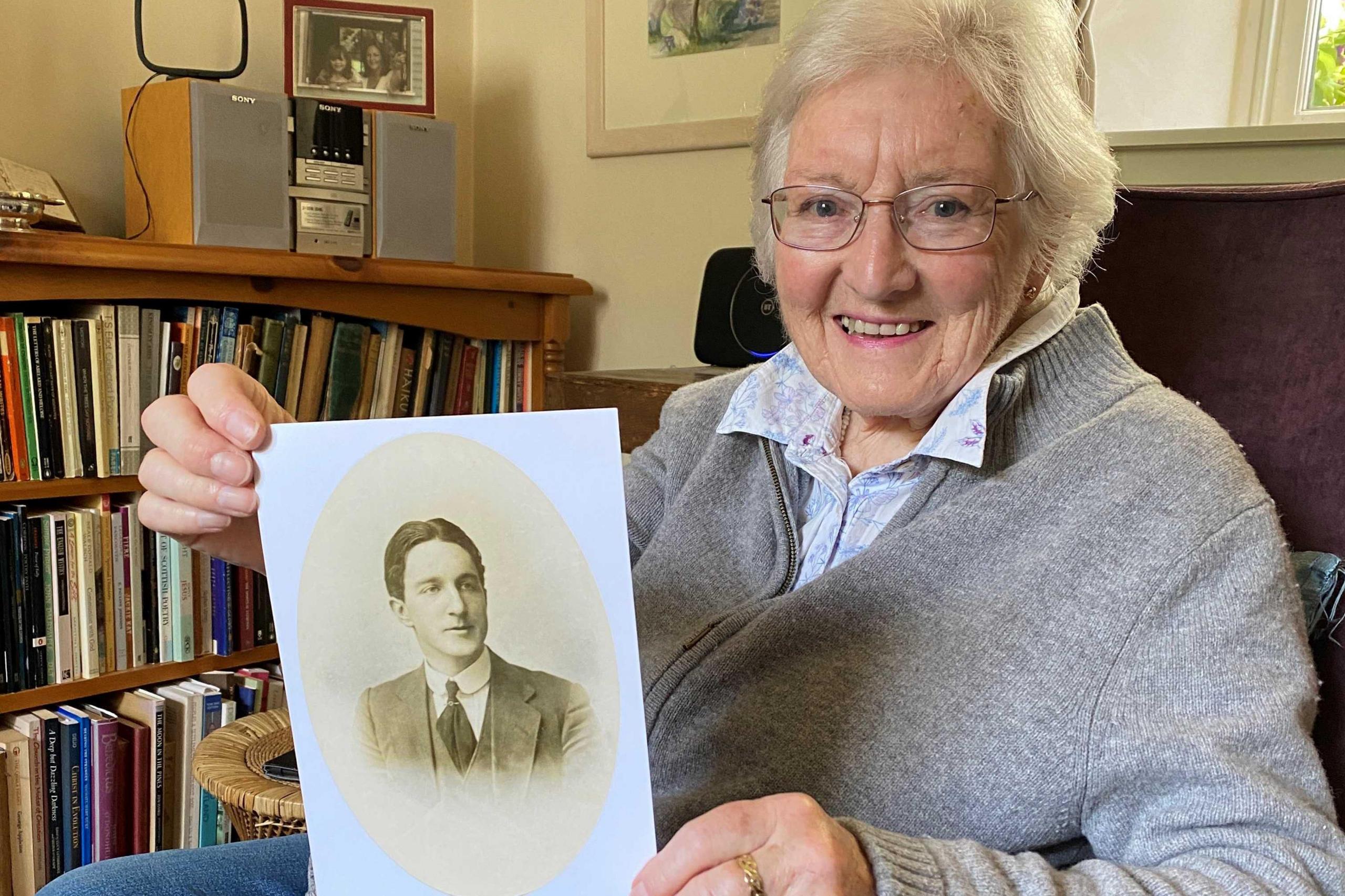 Libby MacRae in an armchair holding a photo of her uncle Donnie MacRae. She is in a living room at her home with bookshelves behind her chair. She is smiling at the camera. She has grey hair and glasses and is wearing a comfortable woollen zip sweater with a shirt underneath and blue jeans. 