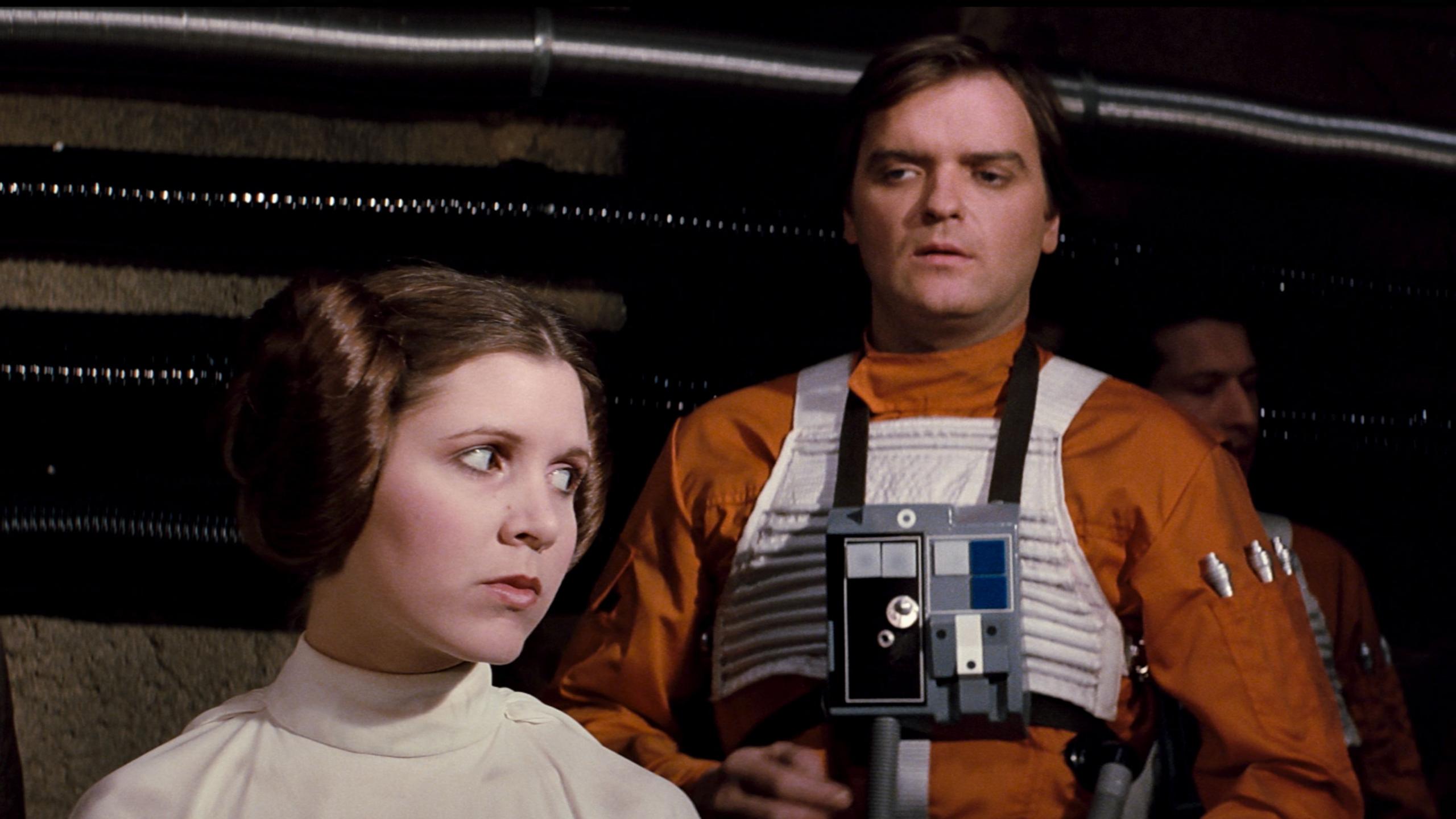 Angus MacInnes is wearing an orange pilot's uniform in a scene from Star Wars Episode IV: A New Hope. He is standing behind Carrie Fisher, playing Princess Leia, who is looking to her left.