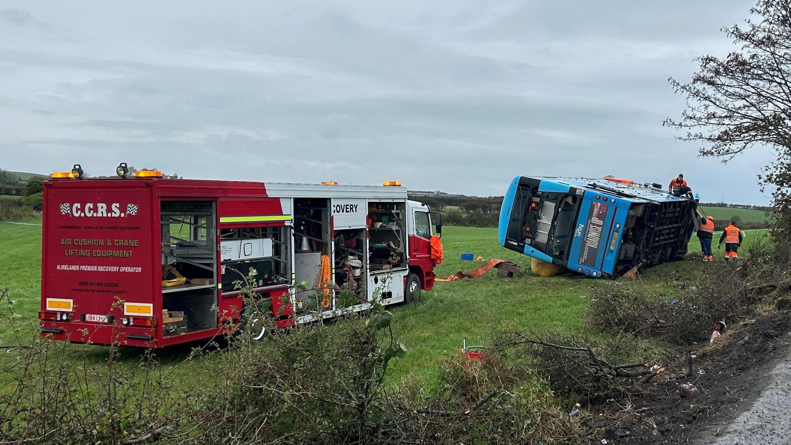 A blue Ulsterbus lies on its side in a field. There are removal workers standing around wearing orange high visibility jackets and red removal truck is parked near the bus in the field. The damaged hedge can be seen in the foreground of the picture.
