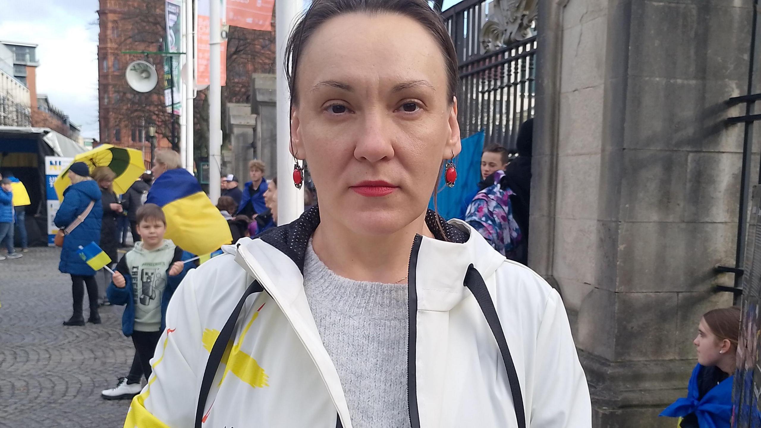 A woman is looking into the camera. She's wearing a white jacket and a grey top and red earrings. People are standing beside her beside gates. The colours of the Ukrainian flag, blue and yellow, are in the background.