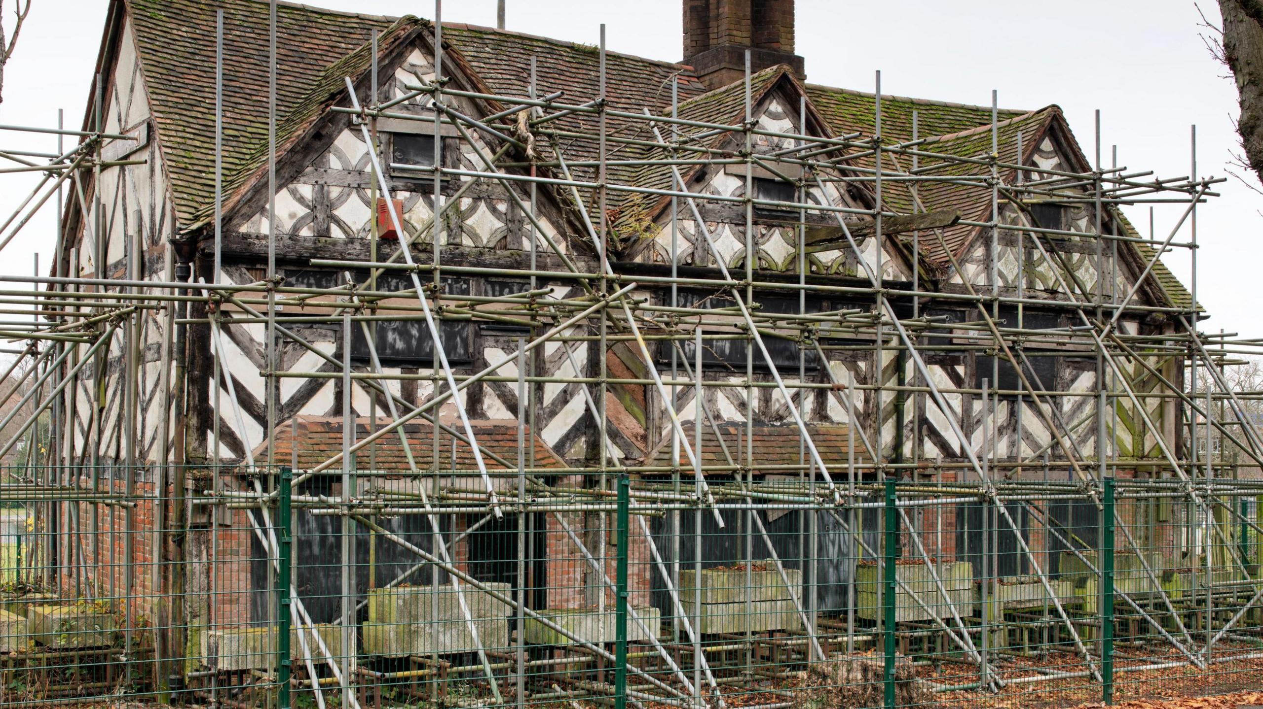 Scaffolding surrounds a timber-framed old pub building, behind a green fence.