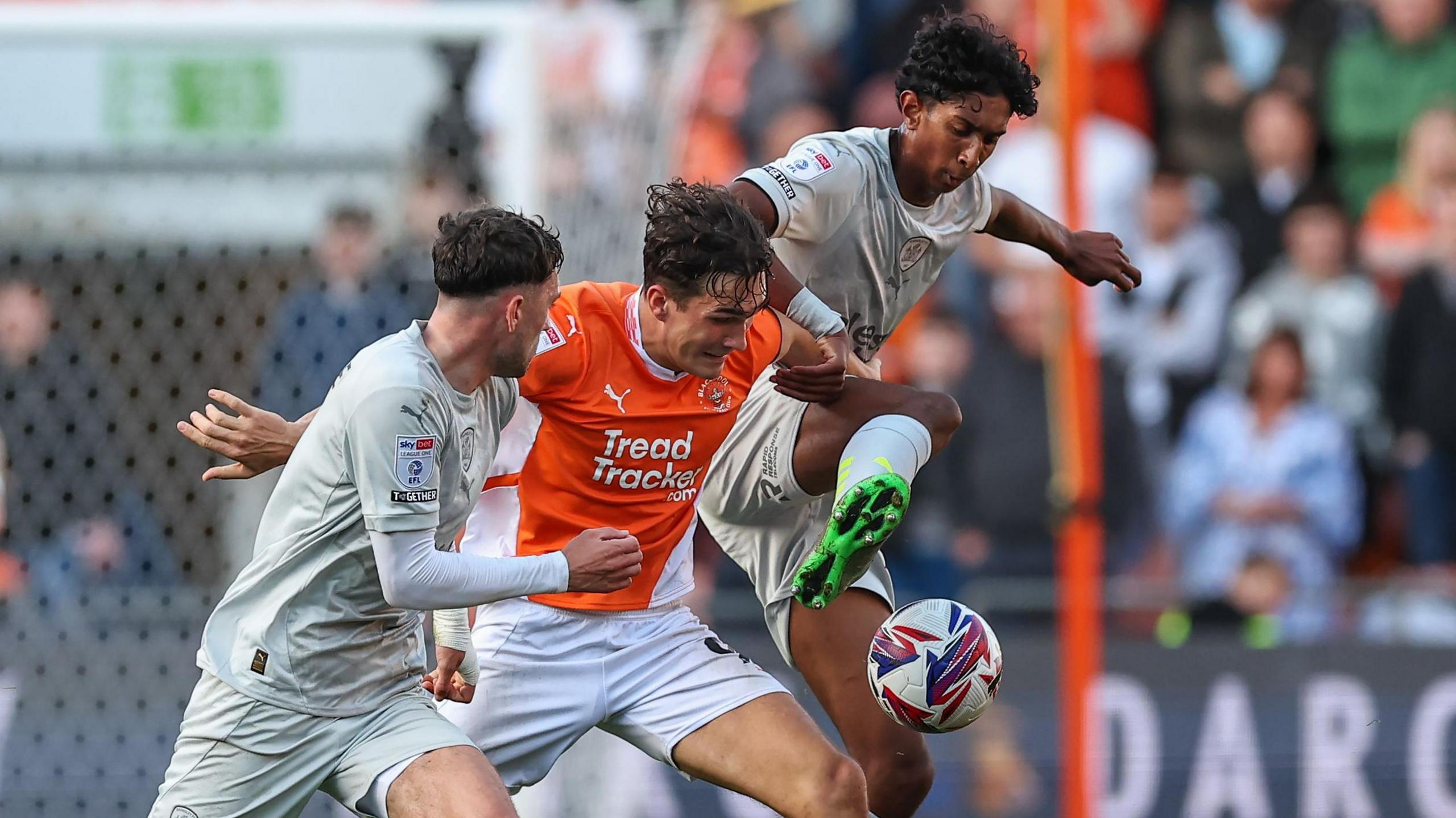 Blackpool's Kyle Joseph battles for possession with Barnsley's Vimal Yoganathan and Corey O'Keeffe during the Sky Bet League One match between Blackpool FC and Barnsley FC at Bloomfield Road on October 19, 2024 in Blackpool. Vimal, in his grey kit, is mid jump as he grapples for the ball. 