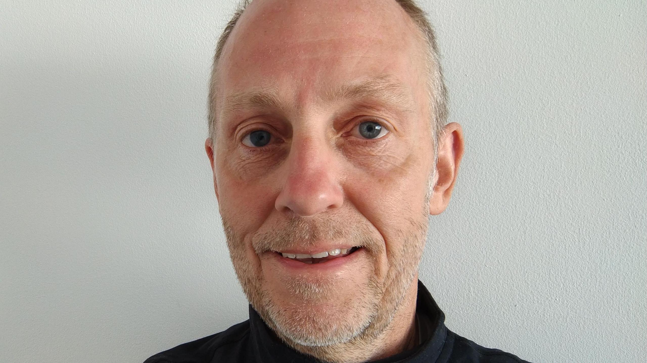 A balding man in a black top staring at the camera, he has light stubble that is mostly grey with flecks of black and the top of a black top is visible on his shoulders