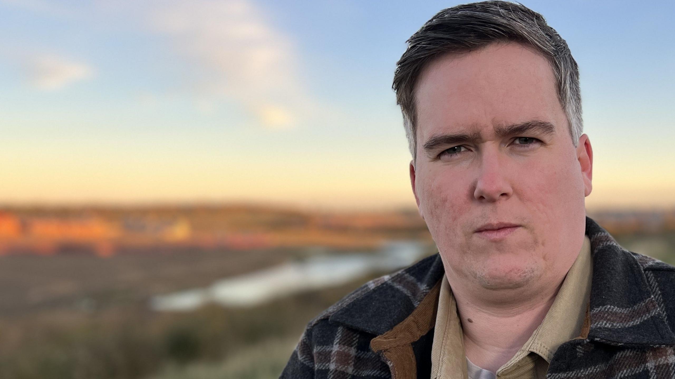 A man with short dark greying hair looks at the camera. He is wearing a beige shirt and a blue checked jacket.