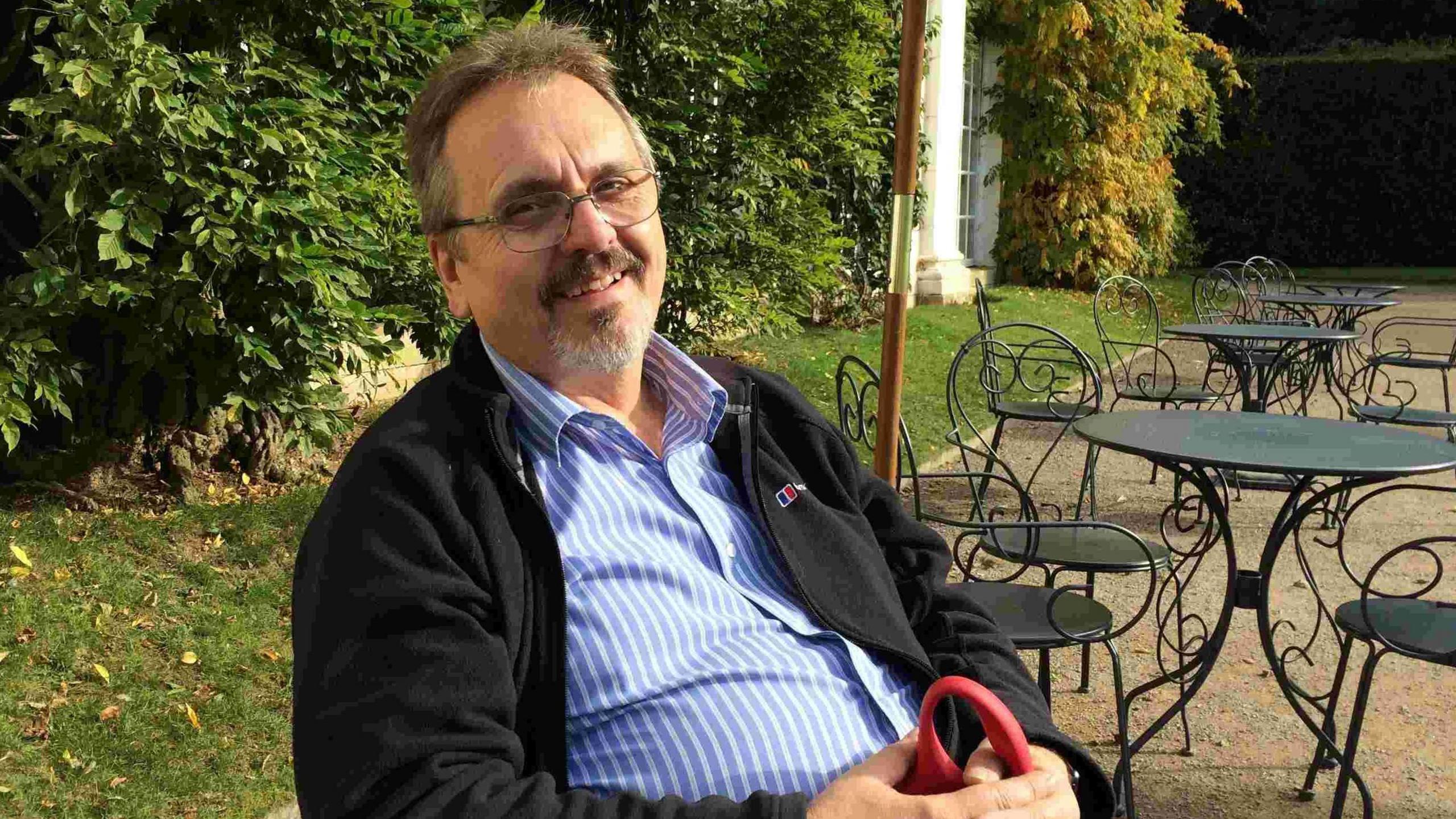 Robert Hewitt wearing a blue and white striped shirt and black jacket sits at a table. He has grey hair and a beard. He is also wearing glasses.