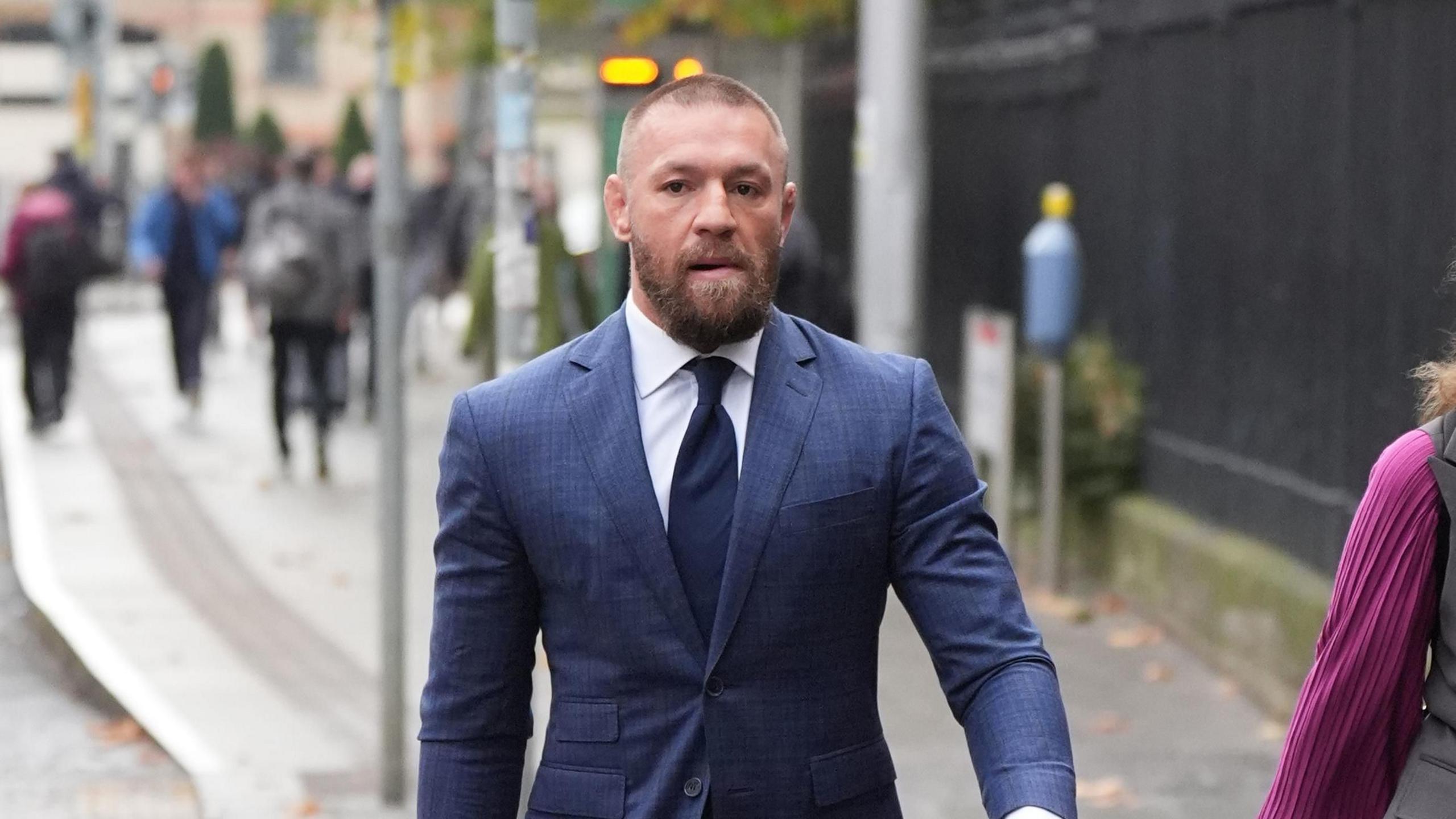 Conor McGregor wearing a navy blazer, white shirt and navy tie. 
He has short fair hair and a beard.