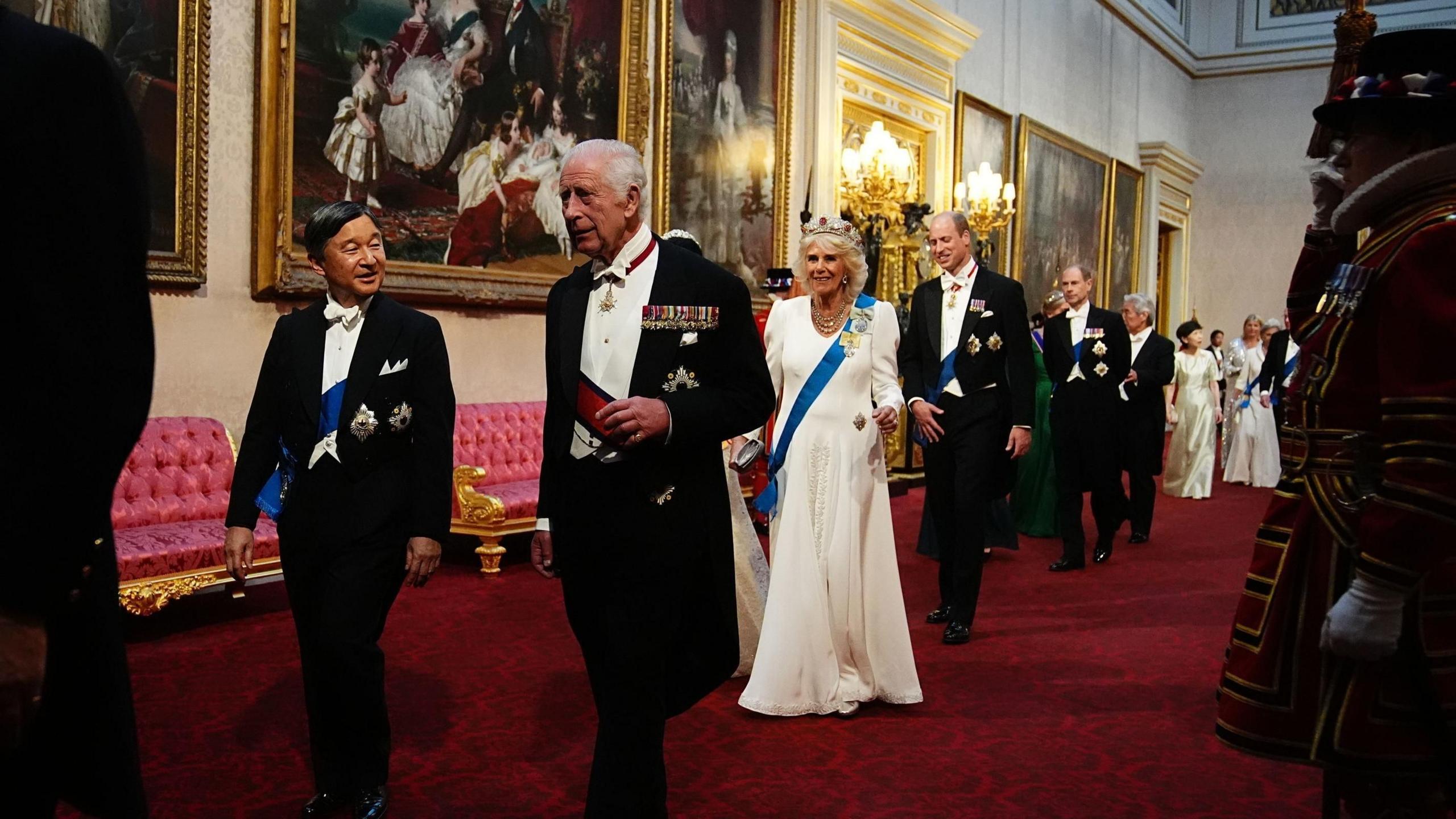 The Emperor and King walking together in Buckingham Palace, followed by the Queen, Empress, Prince William and other Royals. Beefeaters stand at the side