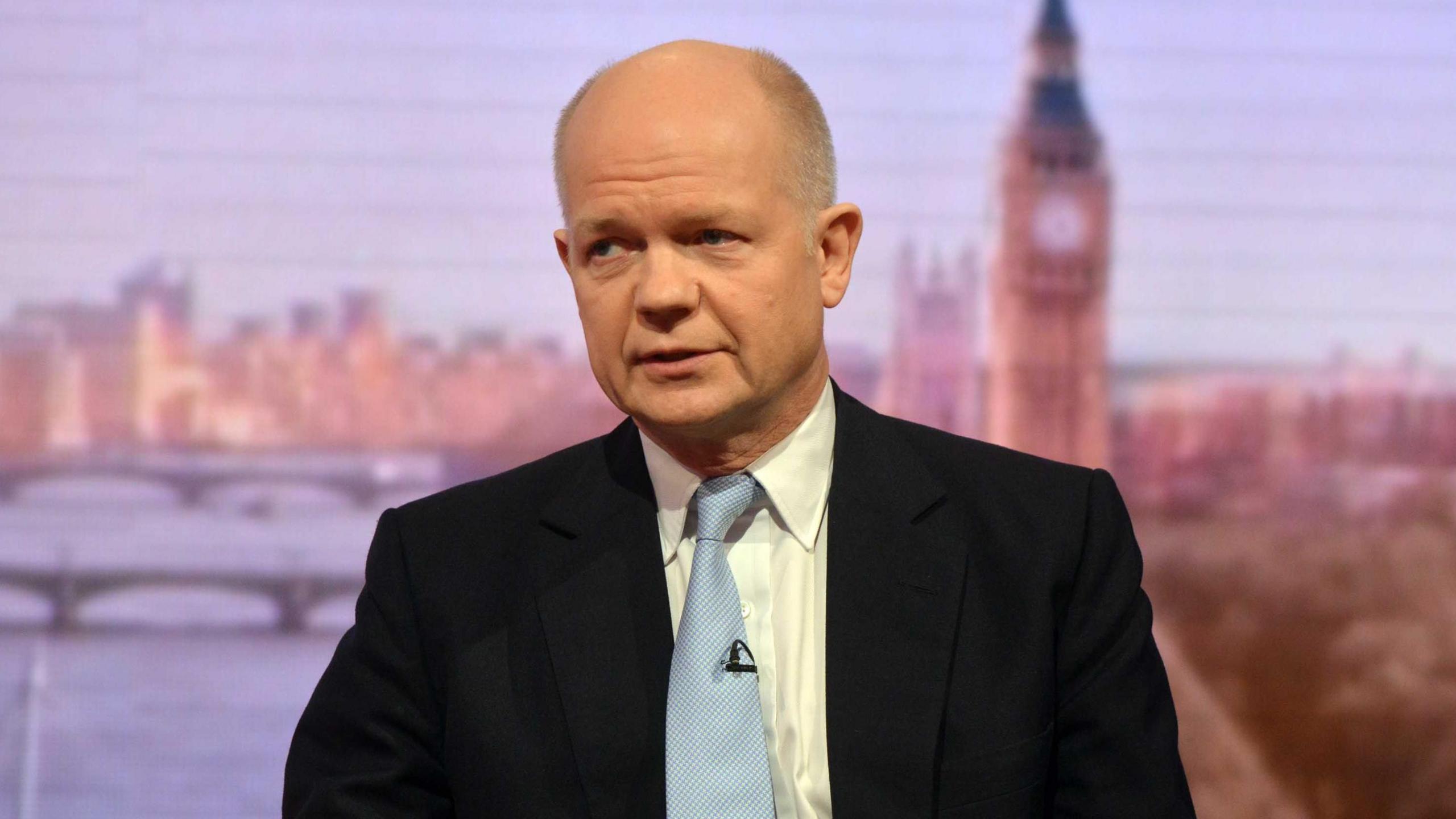 William Hague sat down. He is wearing a black suit, white shirt and light blue tie. He is bald. Behind him is a graphic depiction of the London skyline.