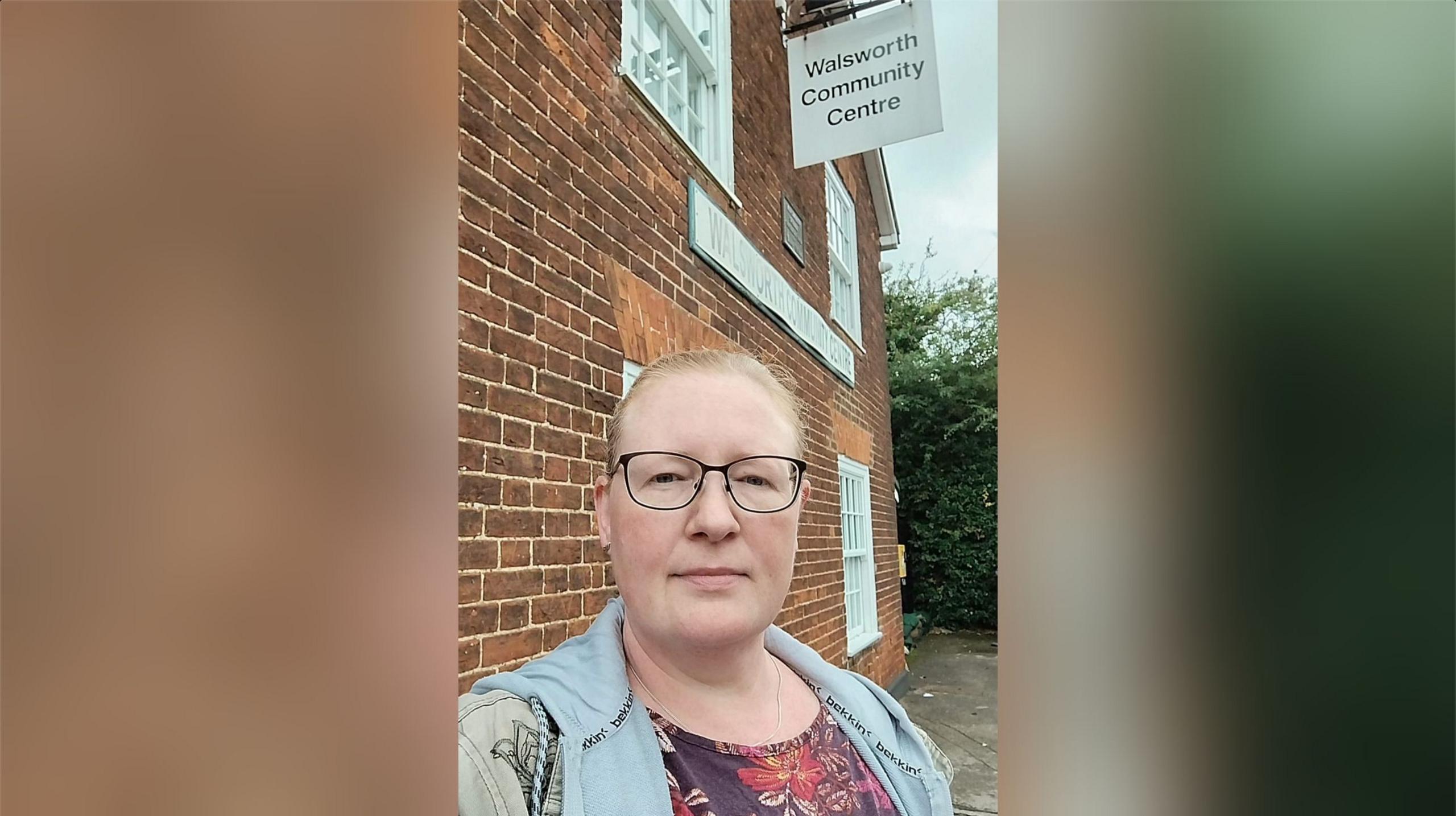 Heike Joachimi with vey short brown hair and glasses, wearing a blue gilet and purple dress outside a two-storey brick building with "Walsworth Community Centre" sign