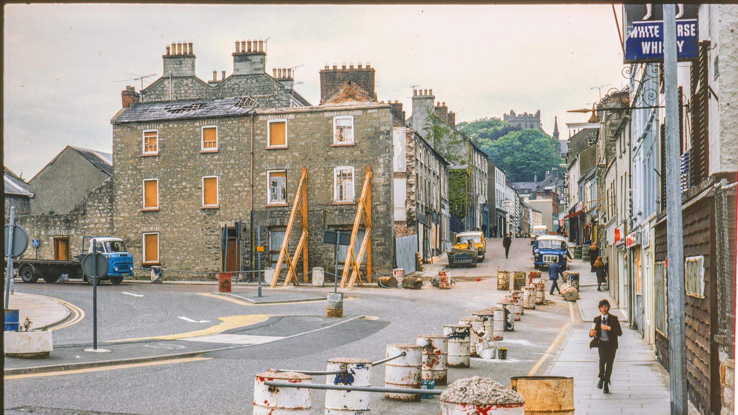 A digitalised photo from a film of a street with oil drums and scaffolding. 
 A boy in Royal School uniform runs towards the camera on the footpath in the foreground which is surrounded by a security barrier made from concrete filled oil drums. The façade of the building on the corner of Scotch Street and Dobbin Street has been removed and the side wall is supported by massive wooden trusses, the roof is partially missing and windows boarded up evidently the result of bomb damage.