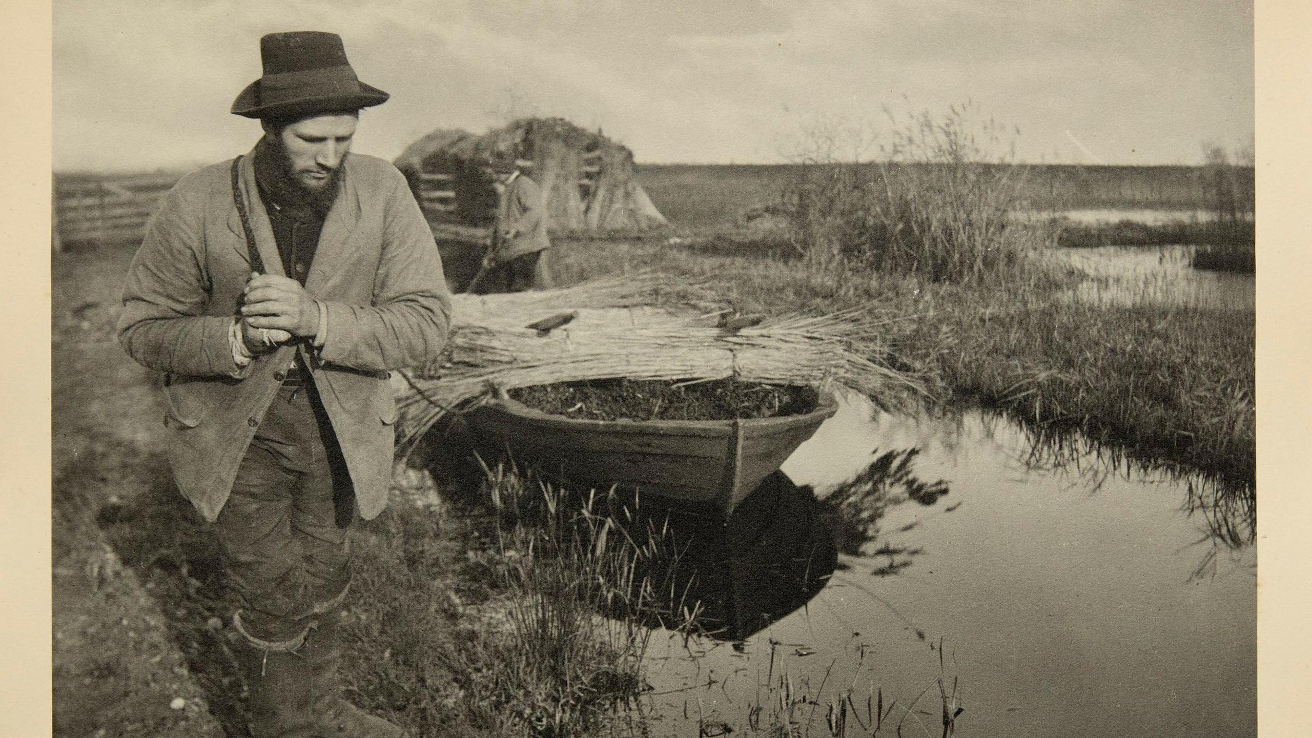 Sepia coloured image of a man on a river bank pulling a small boat filled with reeds.