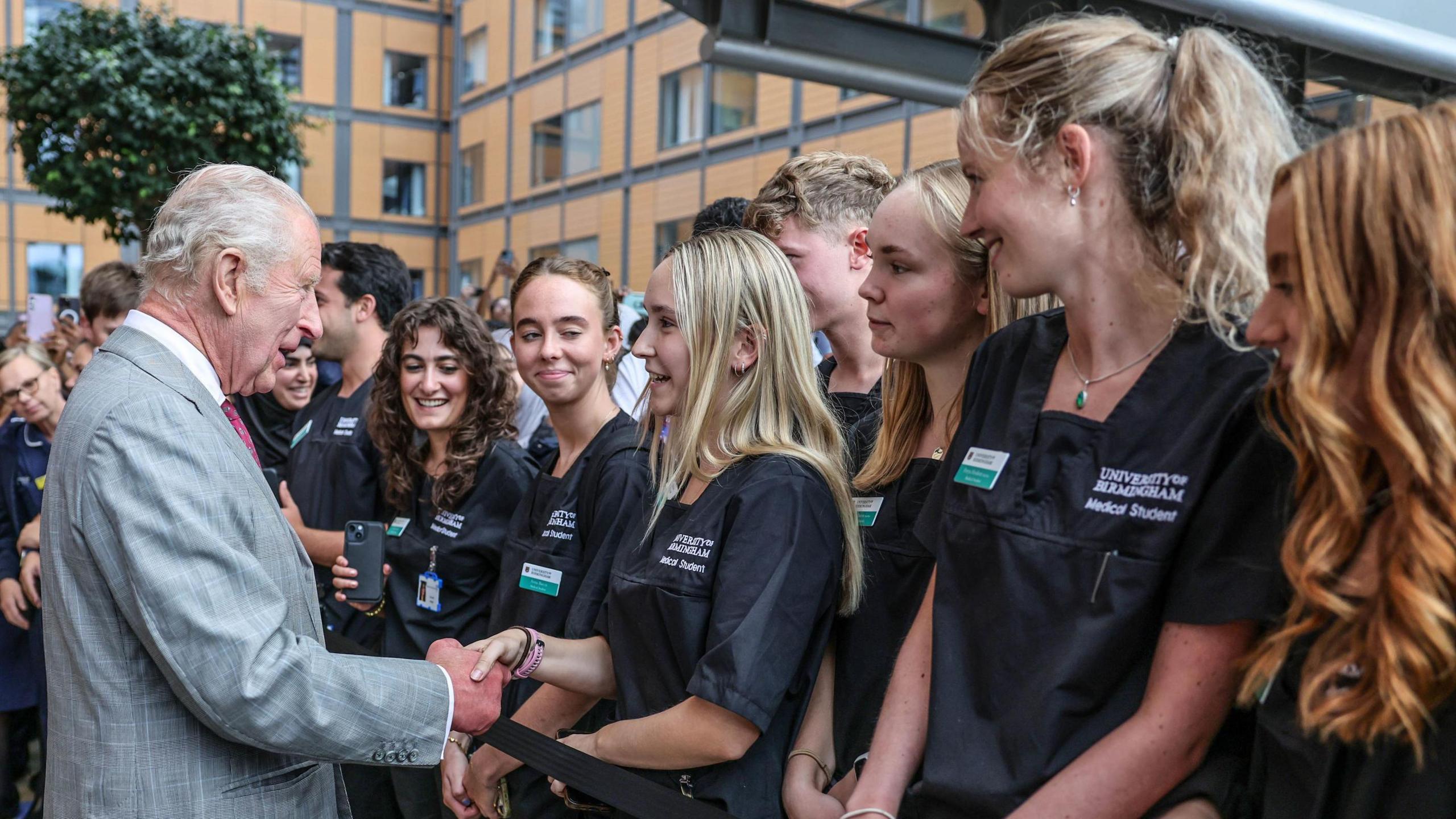 Female medical students in black scrubs stand behind a black tape, facing the King. He is shaking hands with a woman with long blonde hair while crowds of medical staff look on.