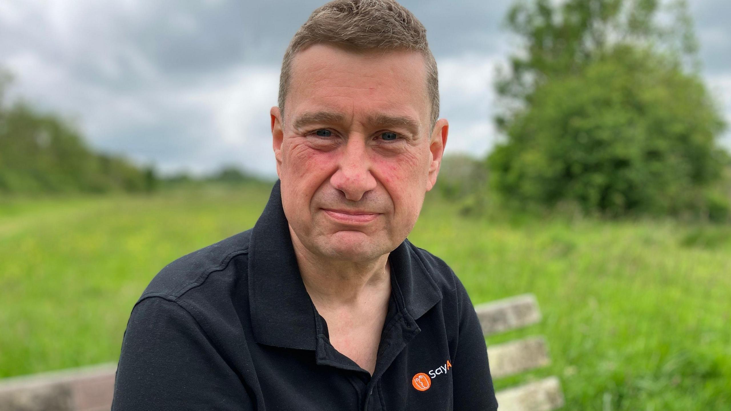 Jez Hodgkinson sits on a wooden park bench in a field, which is blurred in the background, he is looking slightly off camera talking to the reporter and wears a black collared polo shirt with a Say Aphasia logo on the right side of the top