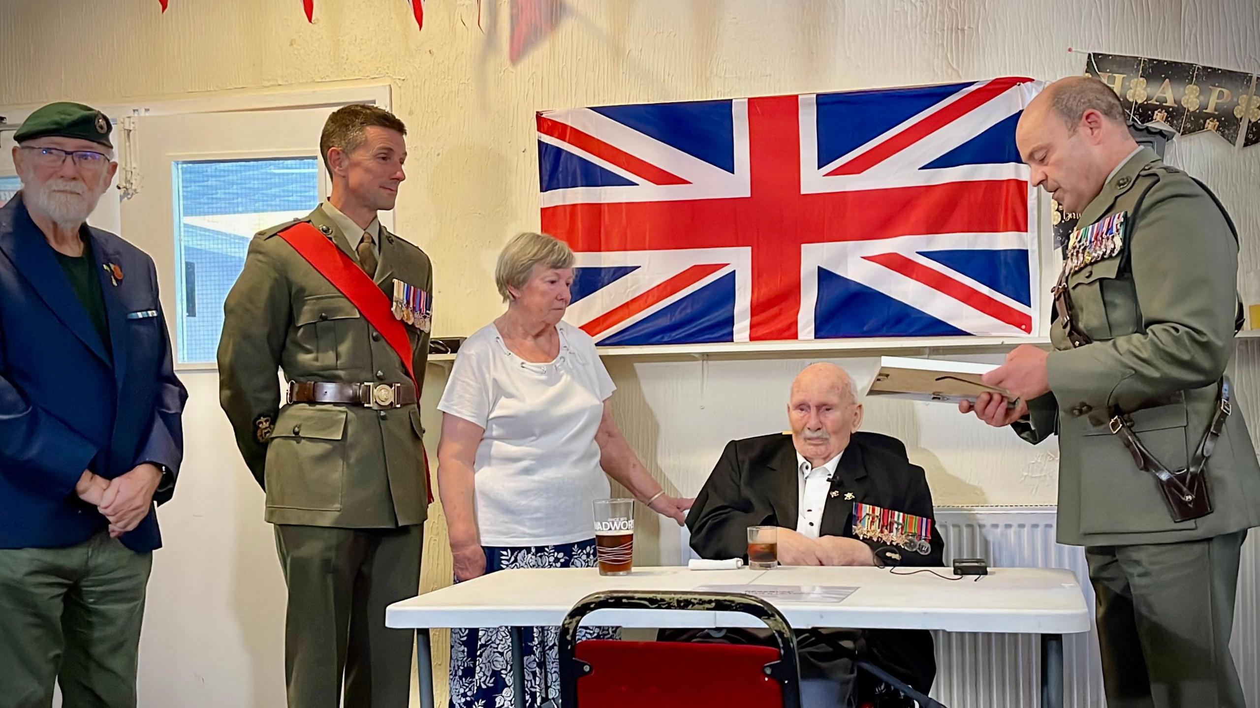 Henry Francis, a 100-year-old World War Two veteran, is seated at a table in front of a large Union Flag. He is surrounded by uniformed military personnel. One stands beside him, reading aloud a letter from Sir Gwyn Jenkins, First Sea Lord, as others look on.